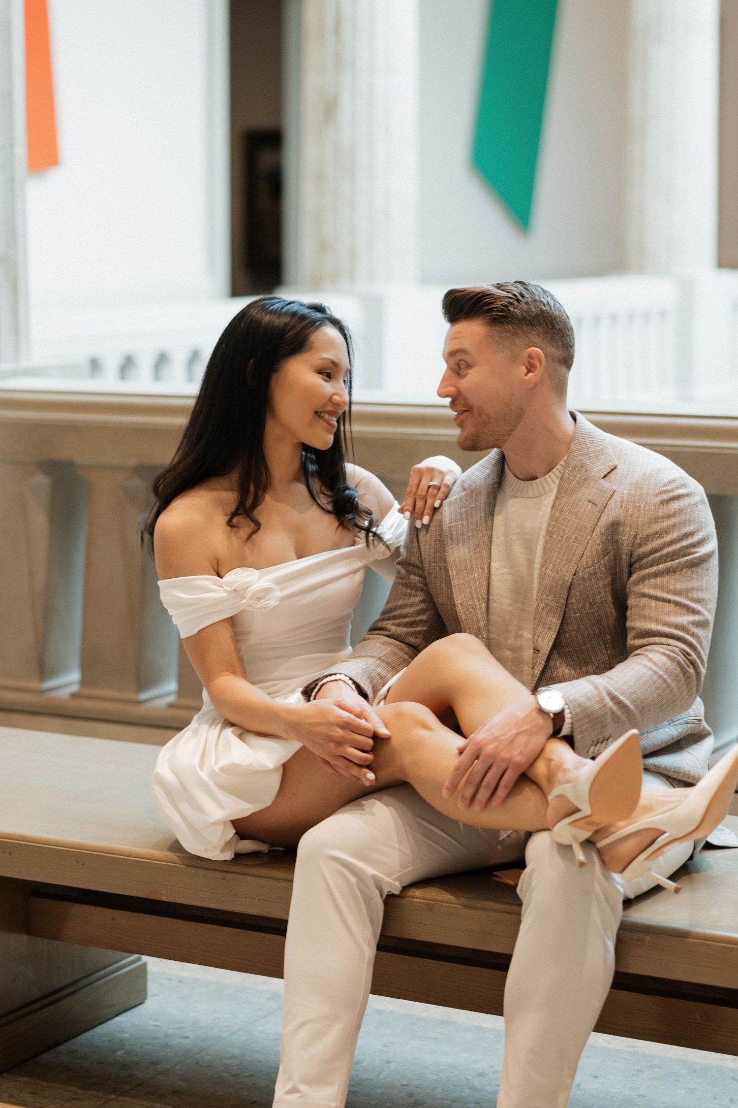 Elegant couple sitting on a bench, woman in white mini dress with legs across man in beige suit, smiling at each other.