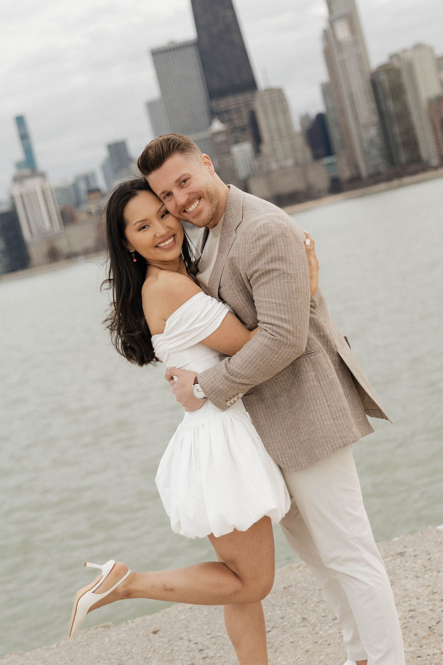 Happy couple hugging near Chicago lakefront, woman in white mini dress and heels, man in tan checkered blazer.