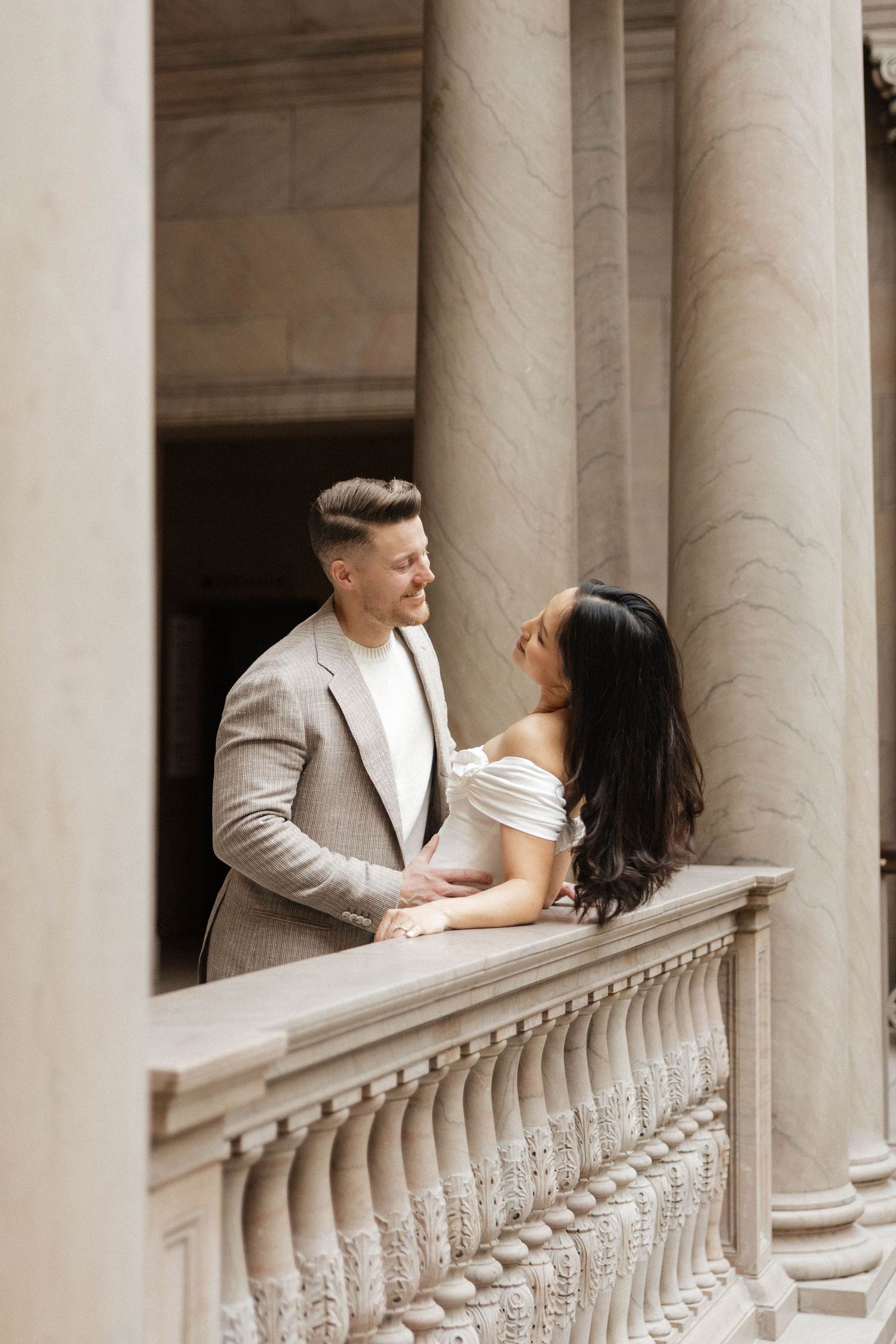 Elegant couple in formal attire sharing a romantic moment by stone balustrade with classical columns.