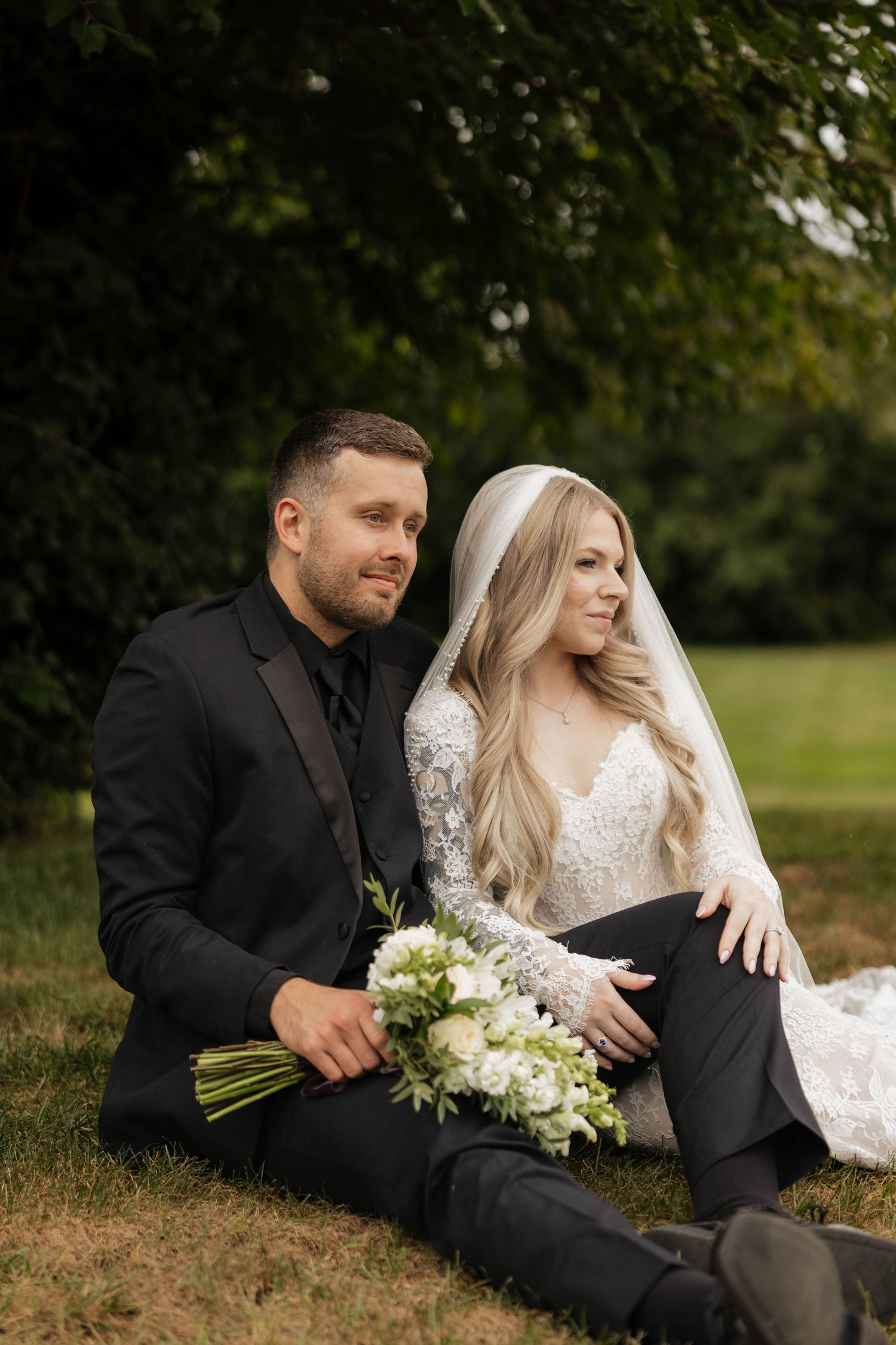 Bride in a lace gown and veil sits beside a groom in a black suit on a grassy lawn, holding a white floral bouquet.