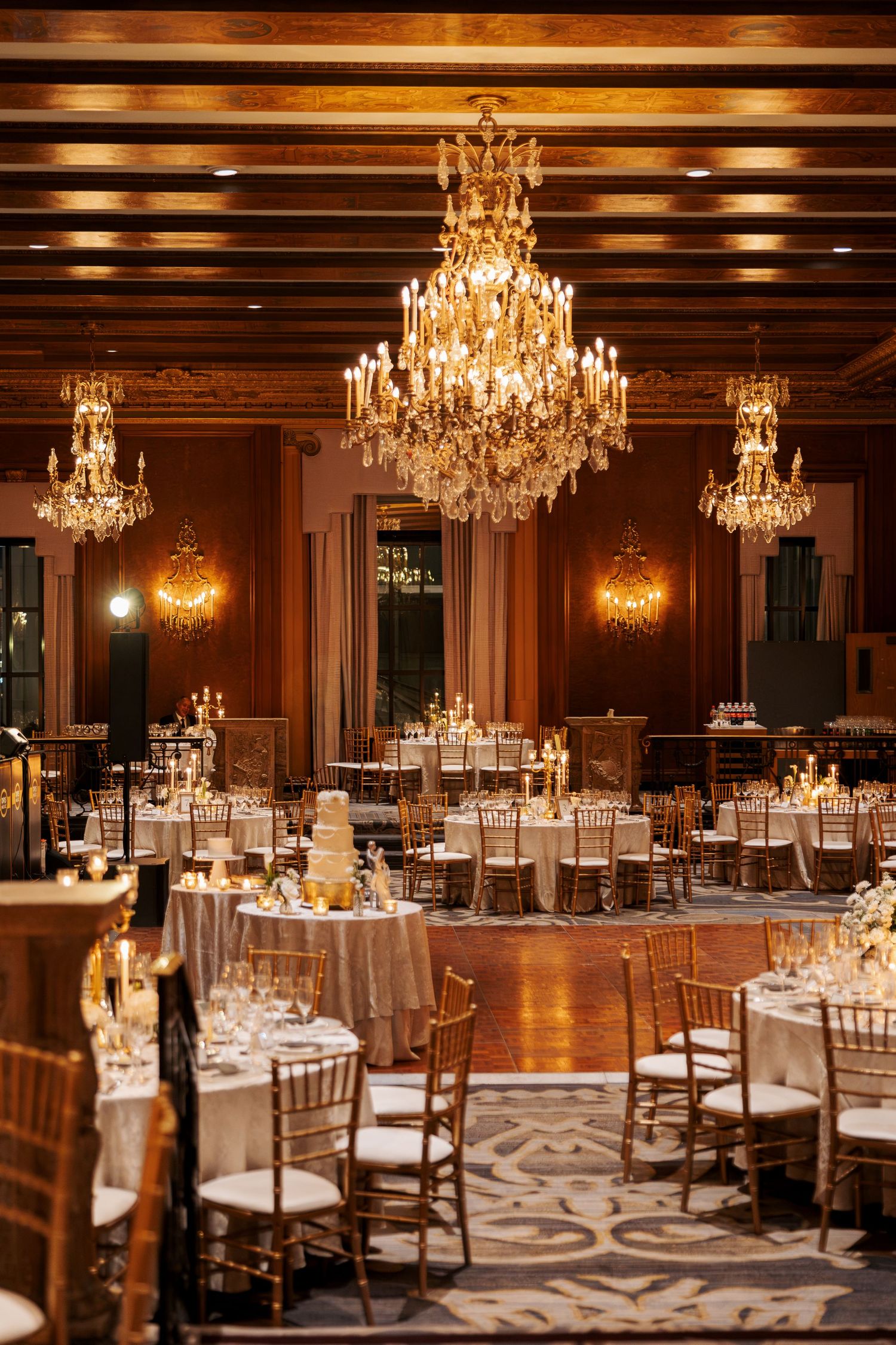 Elegant ballroom wedding reception with crystal chandeliers, white-linen tables, gold chairs and a tiered cake in a warm wood-paneled hall.