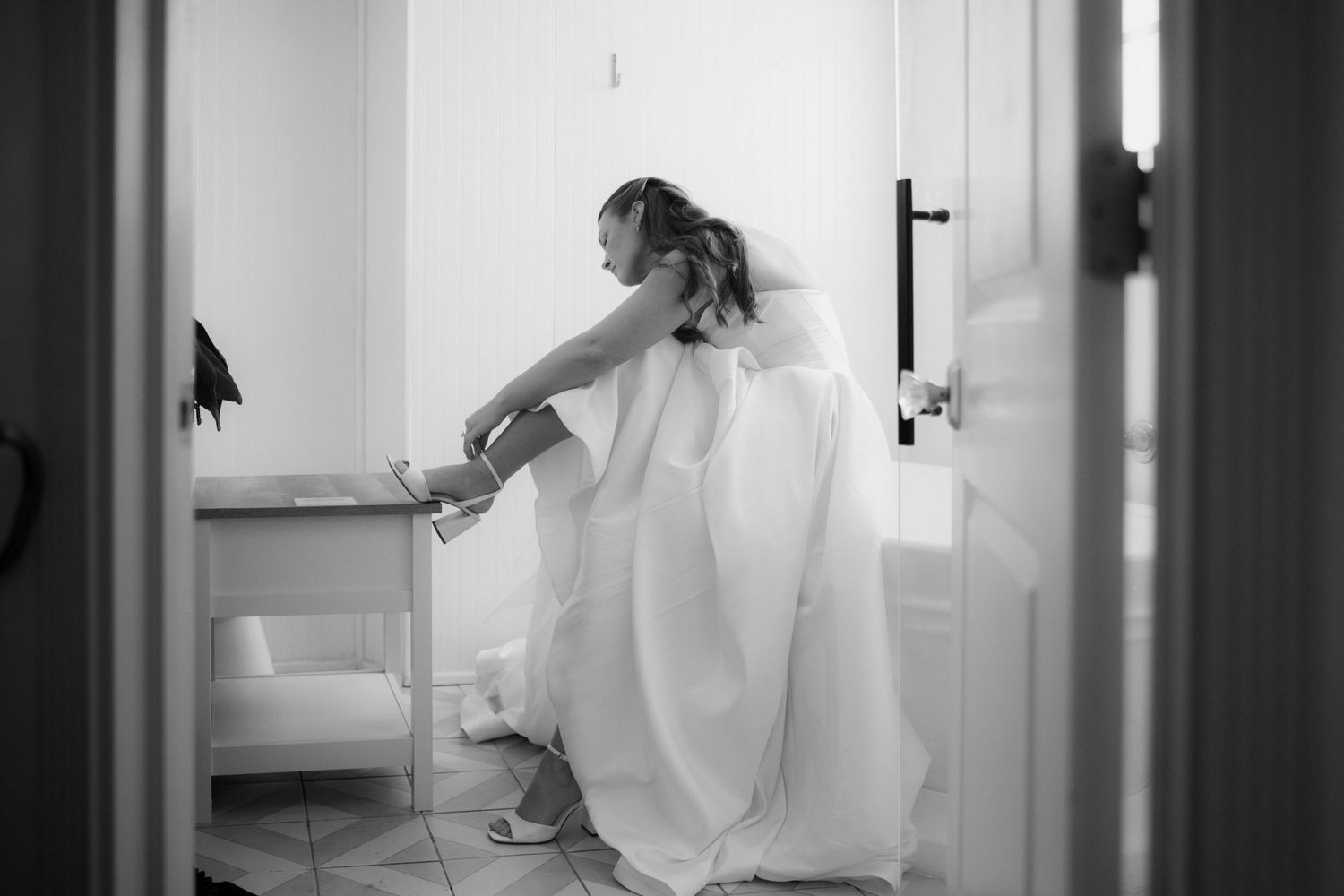 Black and white photo of a bride in a flowing wedding gown sitting in a bright room, adjusting her shoe before the ceremony.