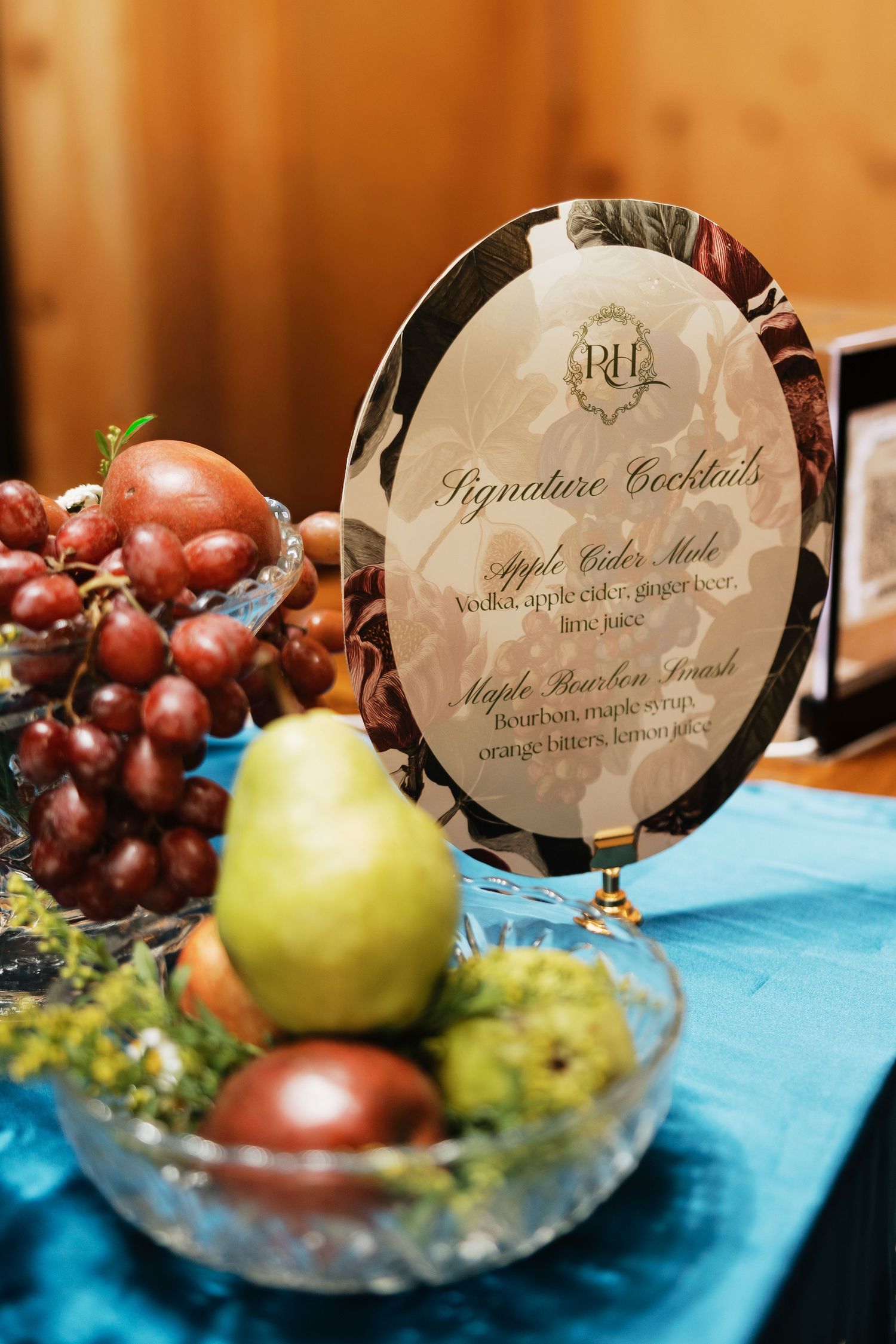 Signature cocktails menu sign with apple cider mule and maple bourbon smash beside a fruit bowl of grapes and pears on a teal table.