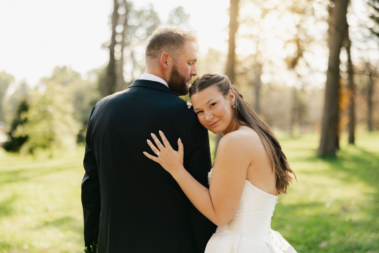 Bride in a white strapless gown rests her head on the shoulder of a groom in a dark suit in a sunlit wooded park.