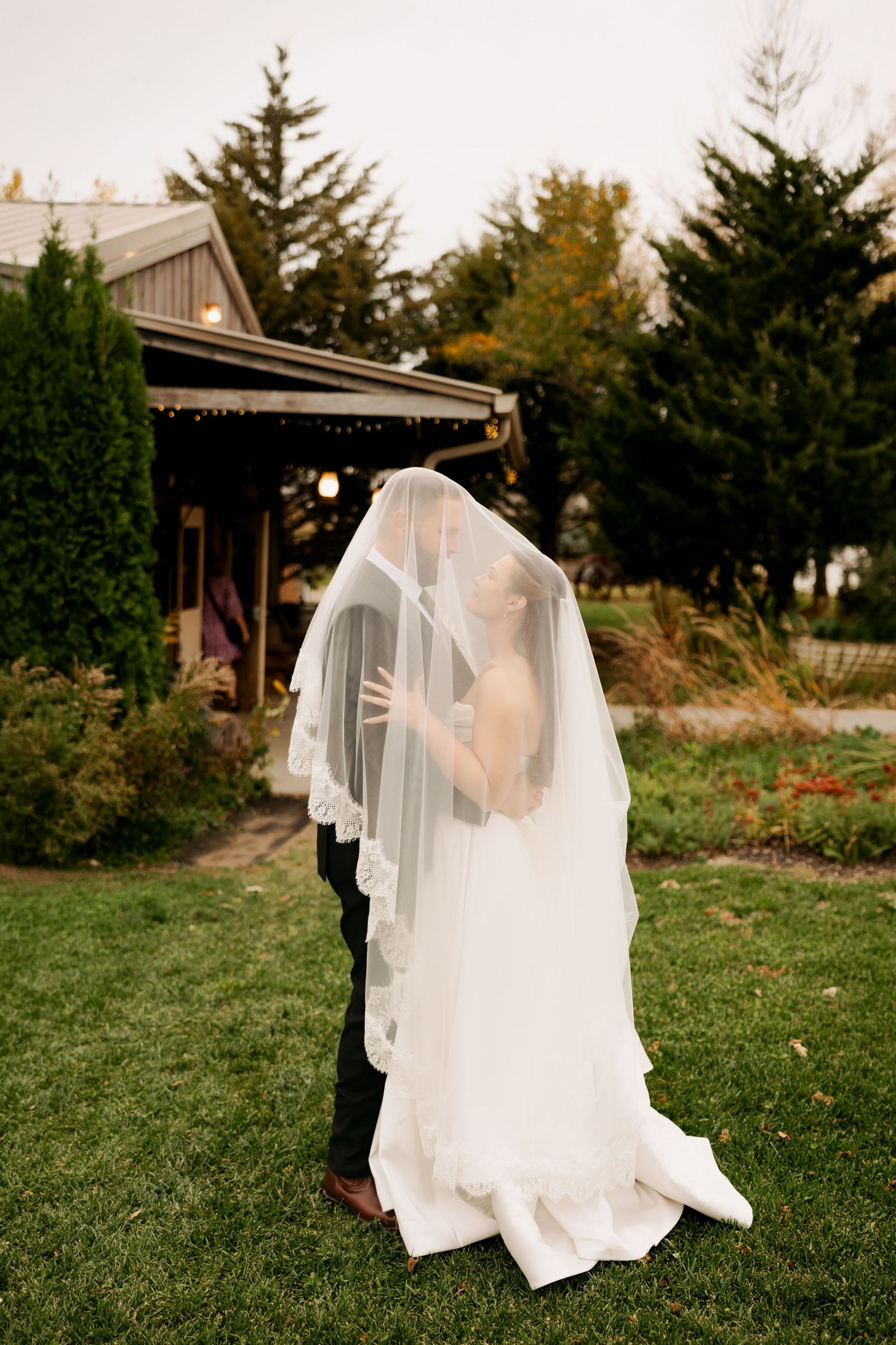 Bride and groom embrace under a long lace-trimmed veil on a green lawn beside a rustic wooden building and evergreen trees.