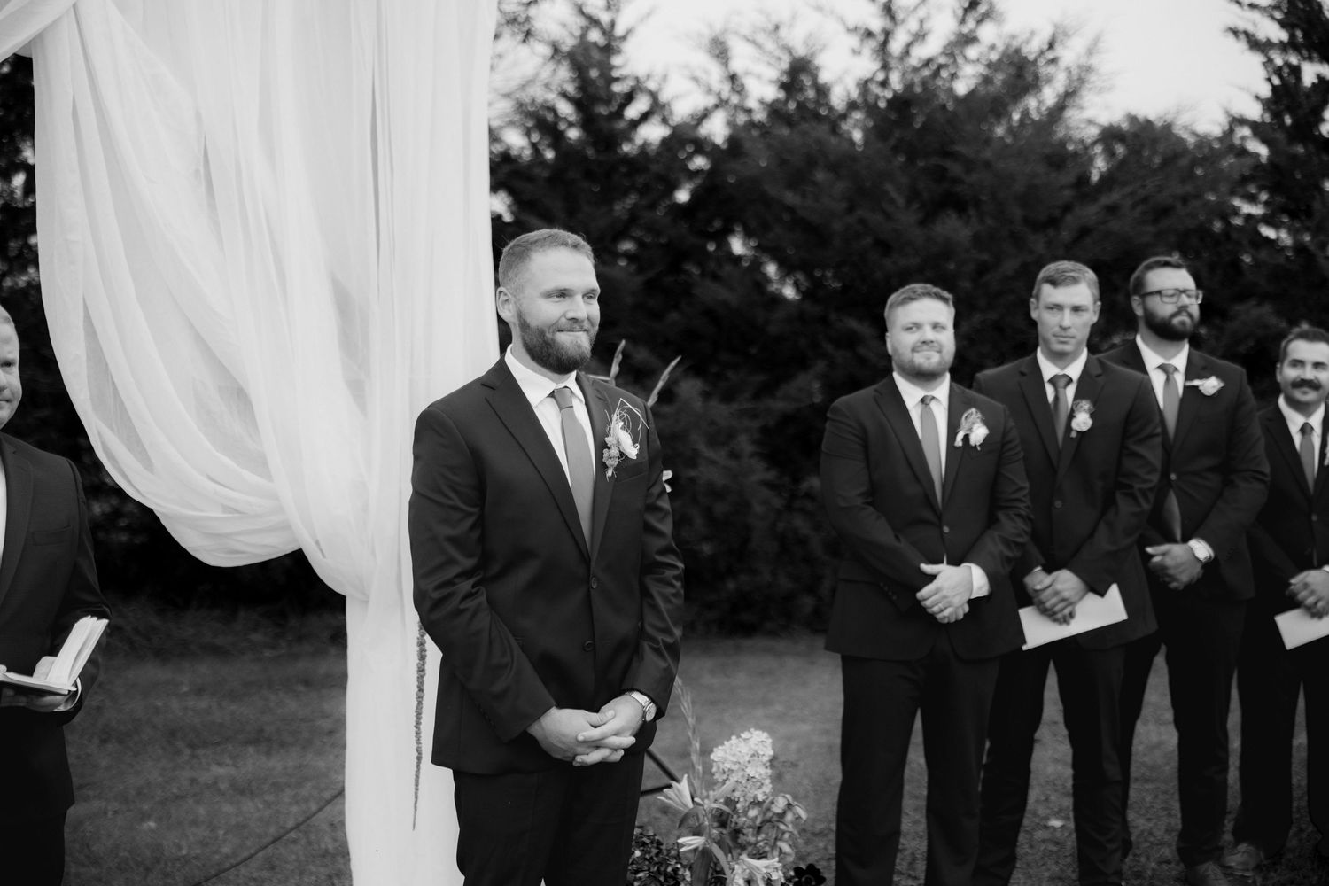 Black and white photo of a smiling groom in a suit standing at an outdoor wedding ceremony with groomsmen lined up behind him.