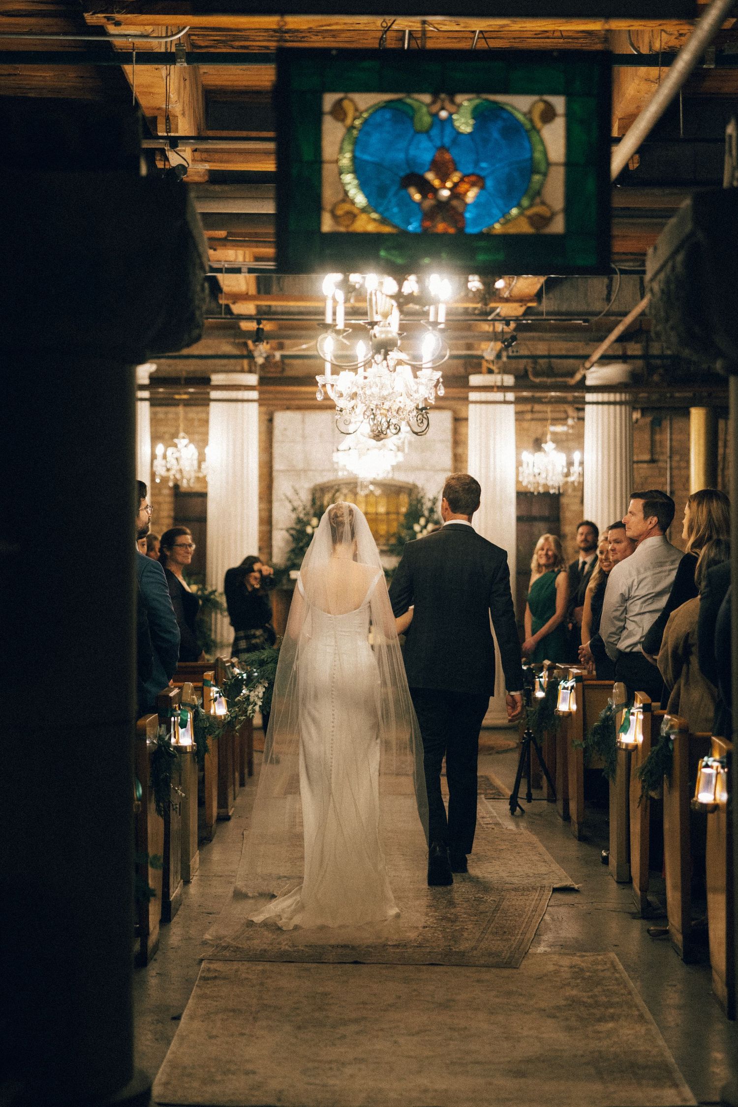 A wedding ceremony in a rustic venue with elegant chandeliers illuminating the aisle and wooden beams overhead.