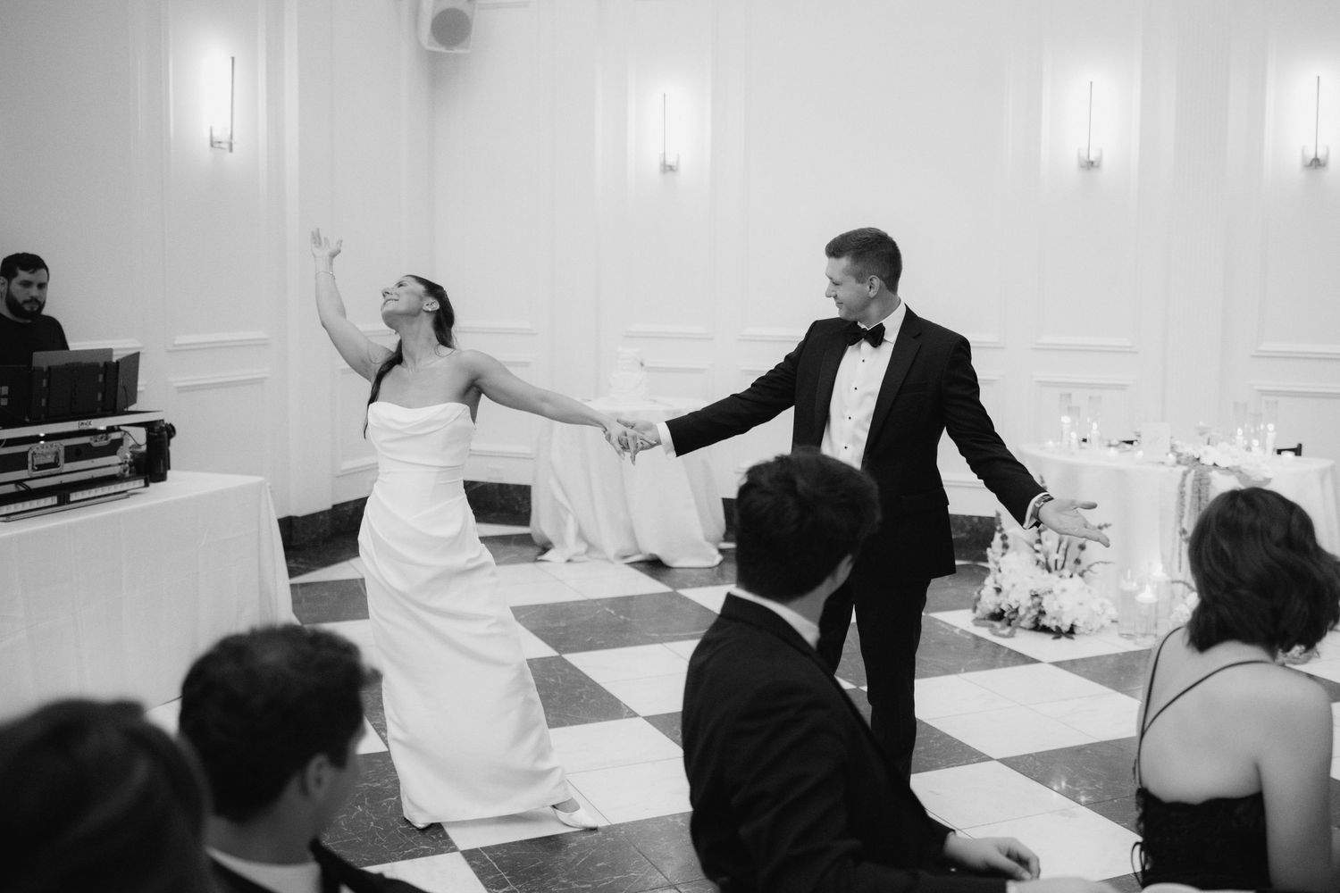 Bride and groom share first dance on checkered floor at elegant wedding reception in black and white photo.