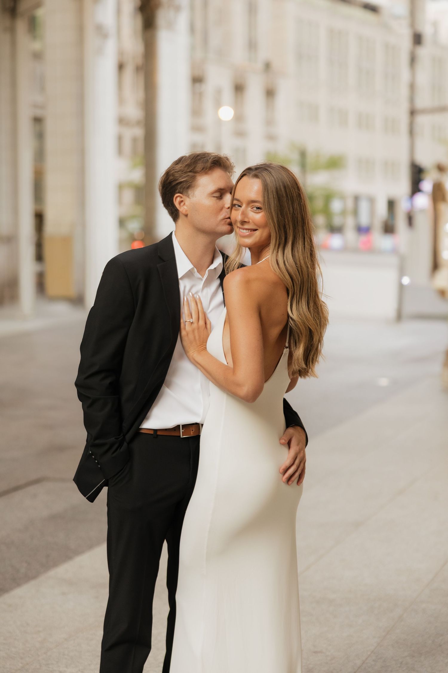 Couple posing for an engagement photo on a city street, the man in a black suit kissing the cheek of a smiling woman in a white dress.