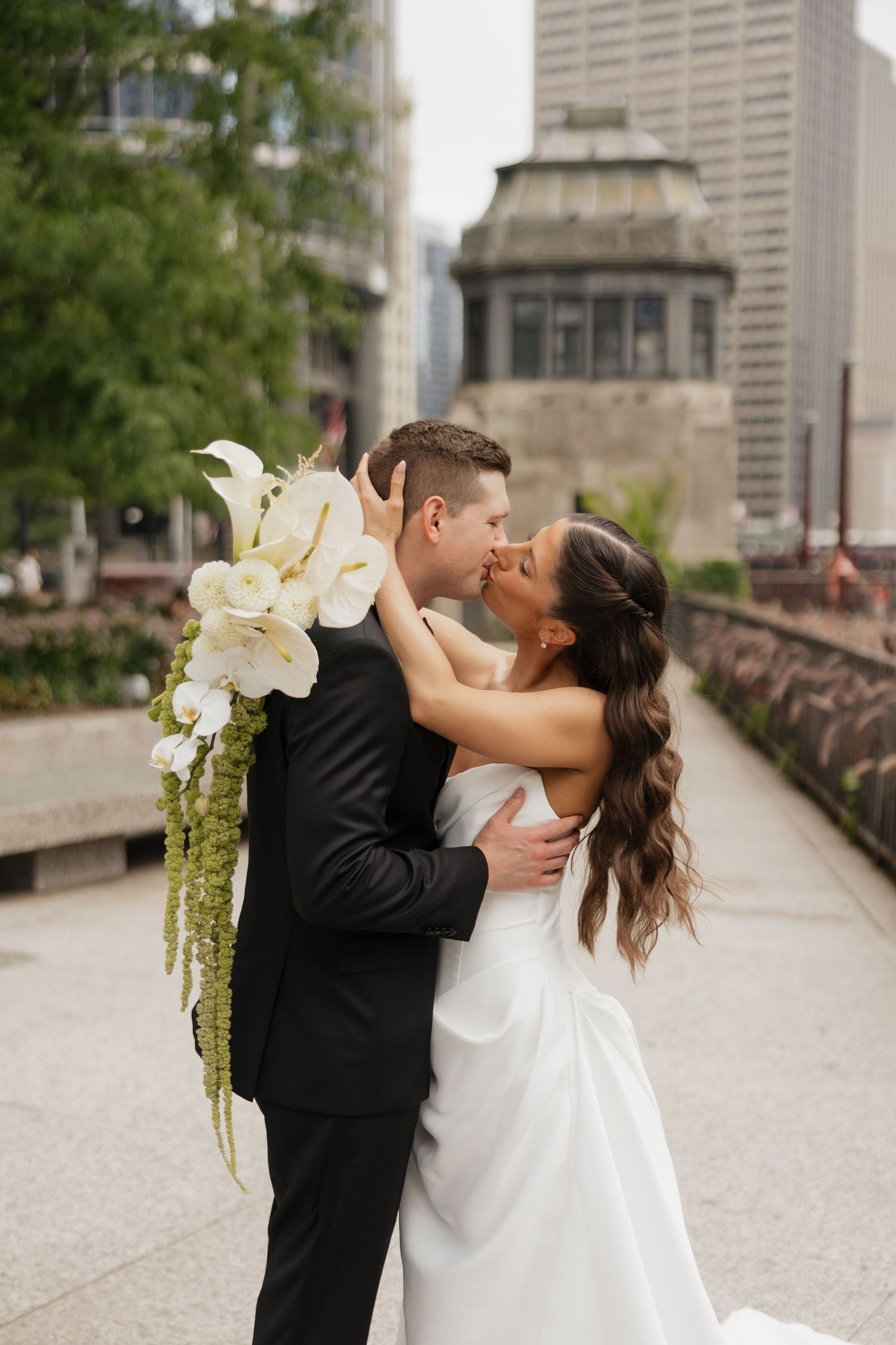 Bride in a white strapless gown and groom in a black tuxedo kiss on a city walkway, holding a cascading white anthurium and green bouquet.