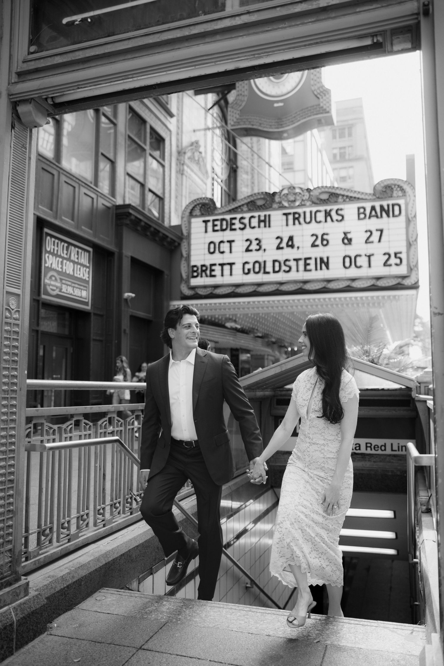 Black and white photo of a couple holding hands descending city subway stairs beneath a theater marquee advertising Tedeschi Trucks Band.