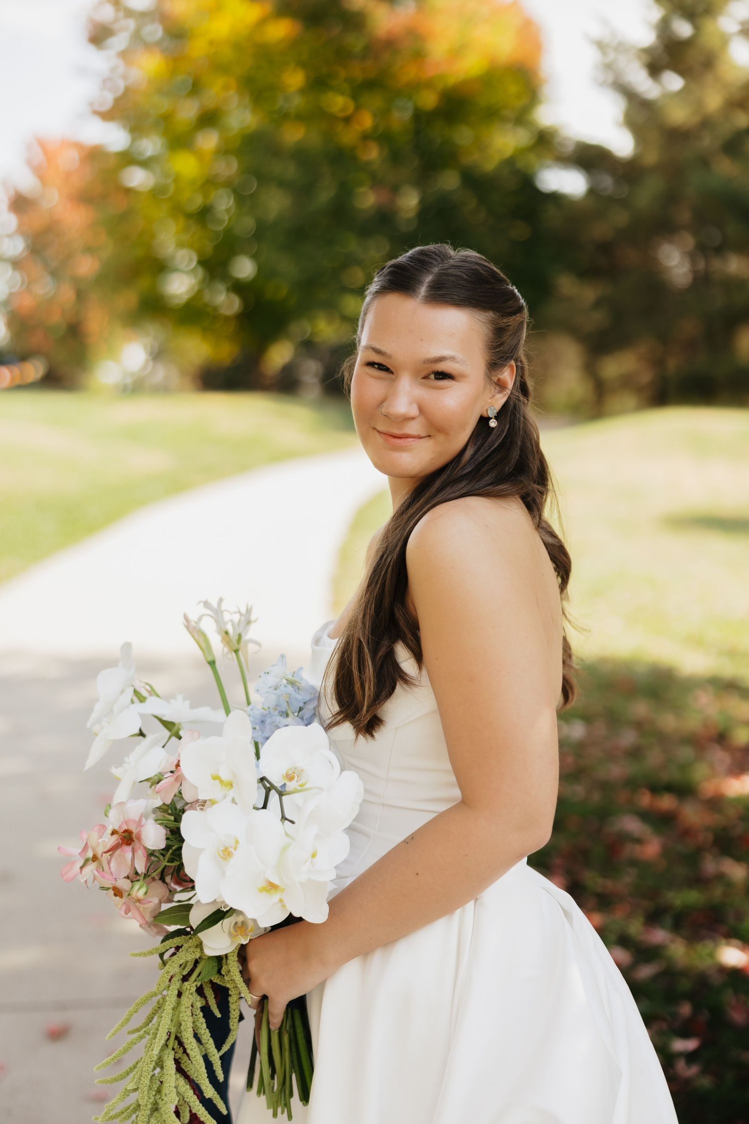Smiling bride in a white strapless gown holding a bouquet of white lilies and greenery on a sunny path lined with autumn trees.