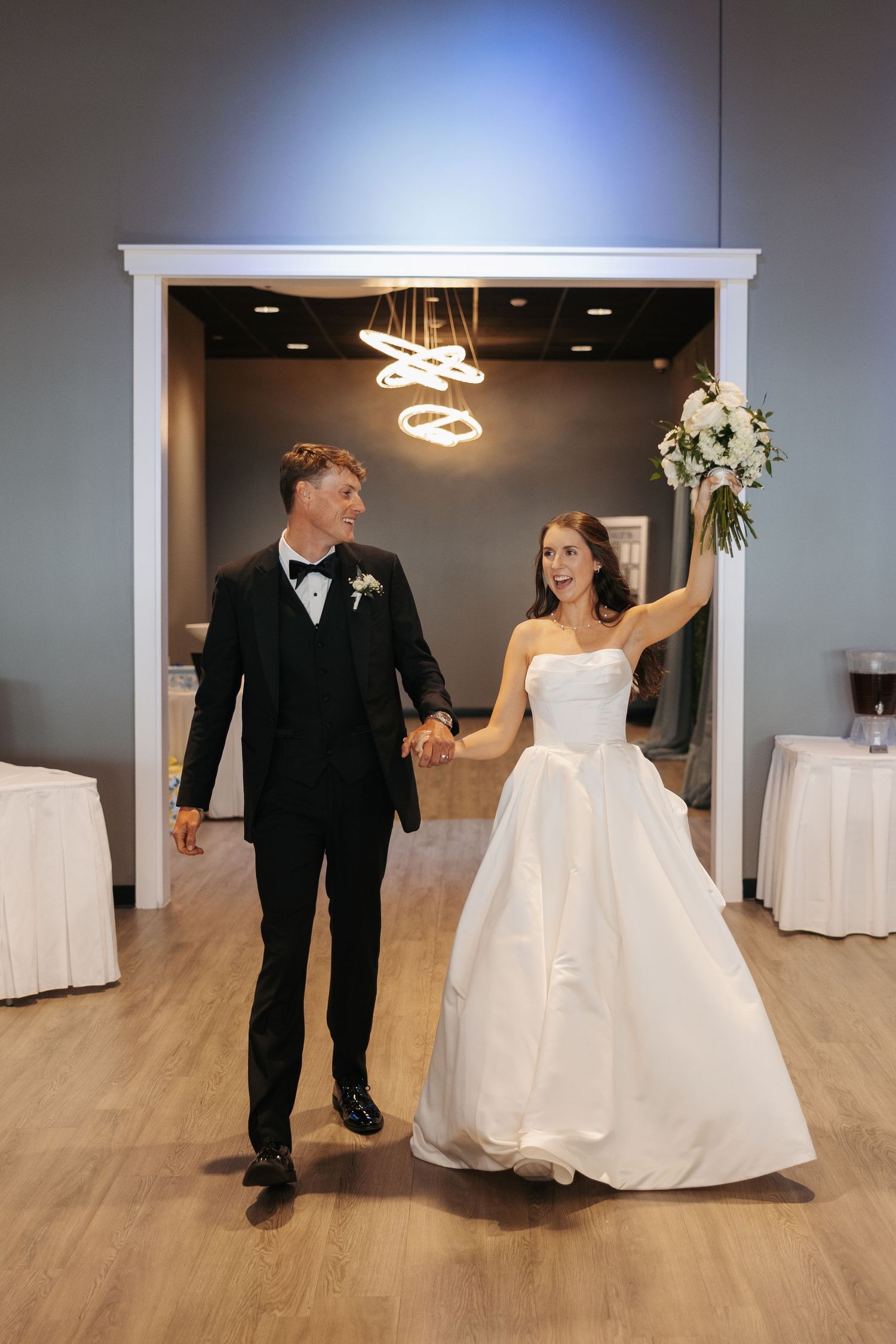 Bride and groom make grand entrance at wedding reception, bride raising bouquet, groom in black tuxedo, smiling joyfully.