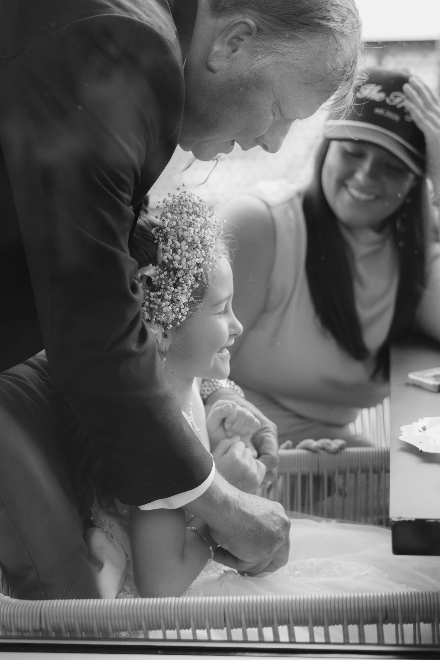 Black and white photo of a child with flower crown smiling at a wedding ceremony with adults nearby.