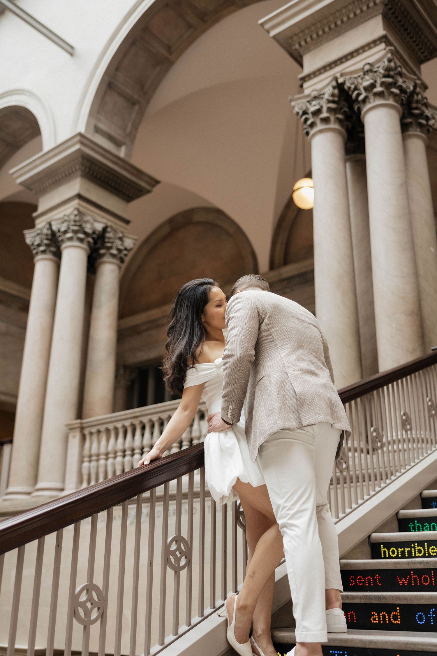 Elegant couple ascending ornate museum staircase with towering columns and arched ceilings in background.