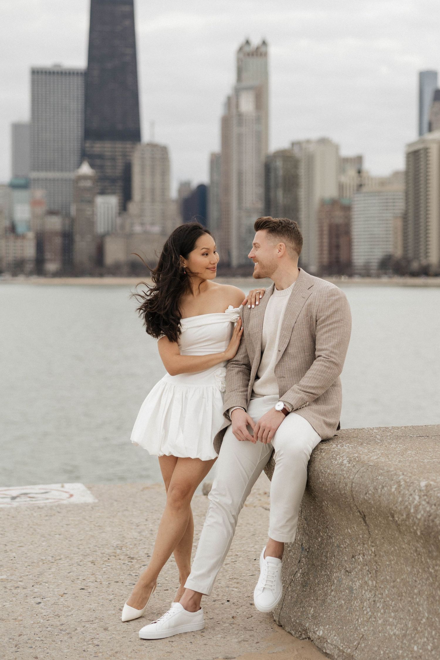 Couple posing romantically by Chicago lakefront with city skyline backdrop, woman in white mini dress, man in beige suit.