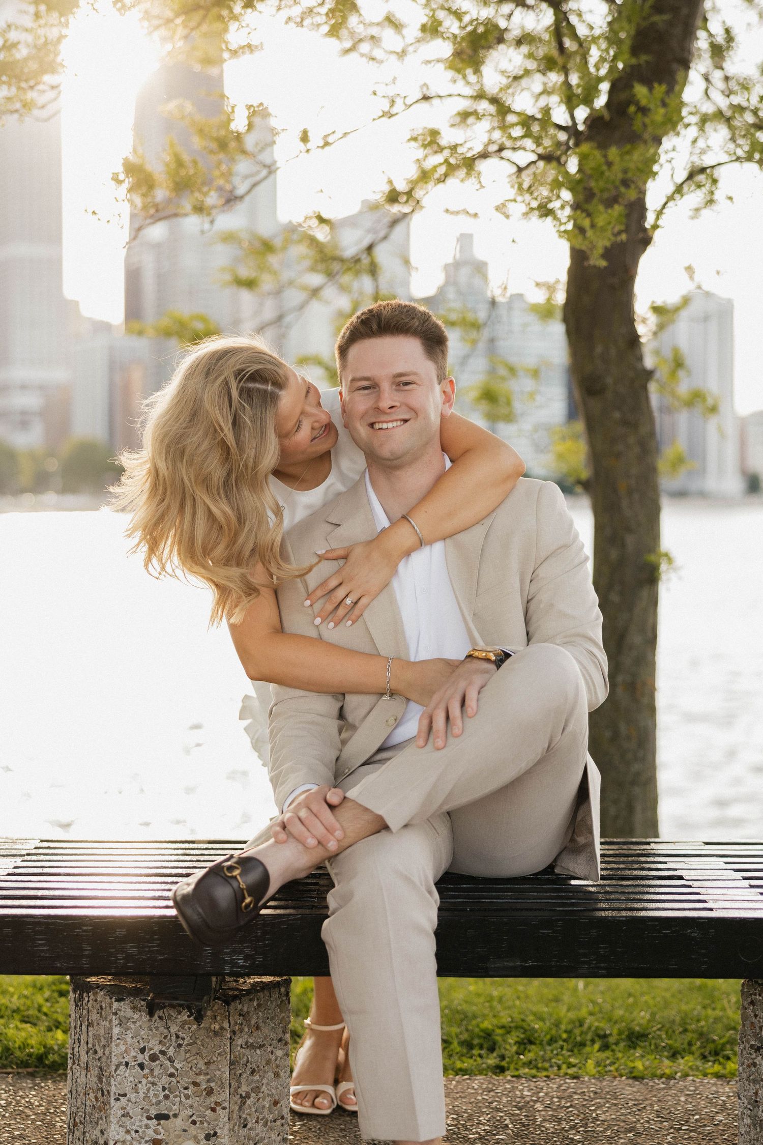 Couple embracing on a park bench by the waterfront at golden hour, man in beige suit, woman in white dress.