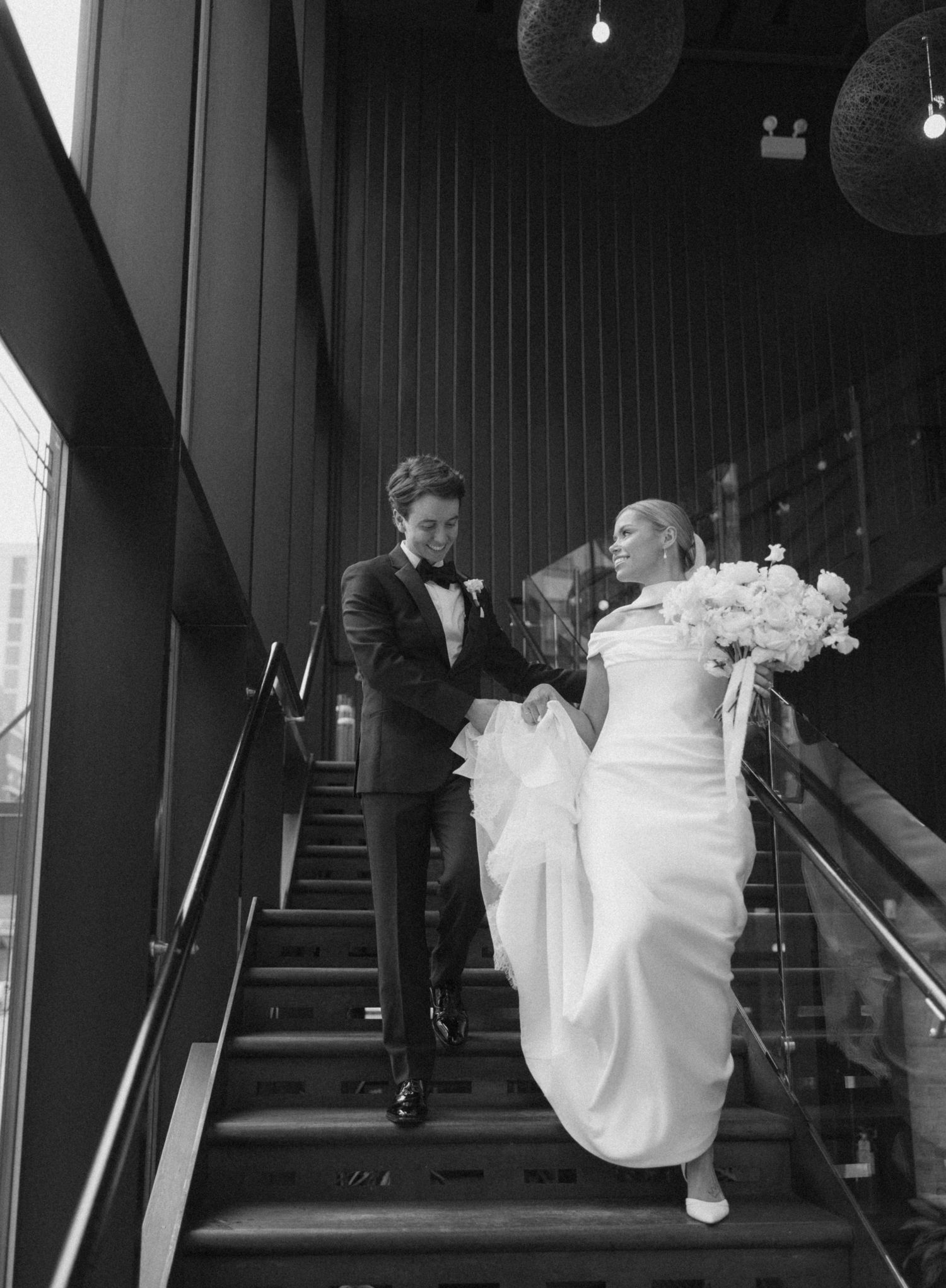 Black and white photo of a bride and groom descending a modern staircase, bride holding white bouquet.