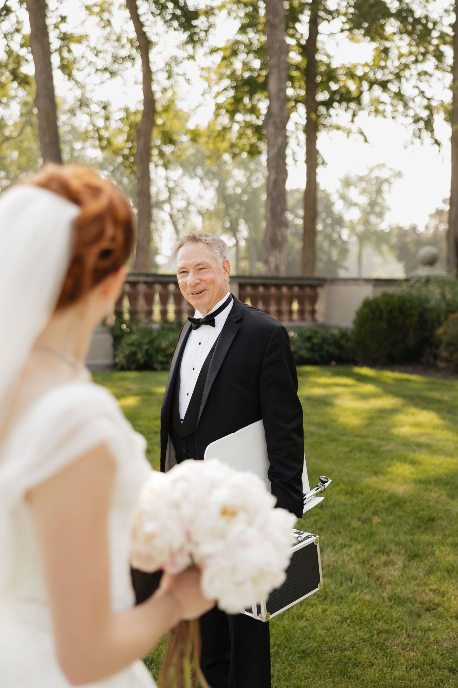 Smiling officiant in tuxedo holds wedding materials while greeting bride with white bouquet on green lawn.