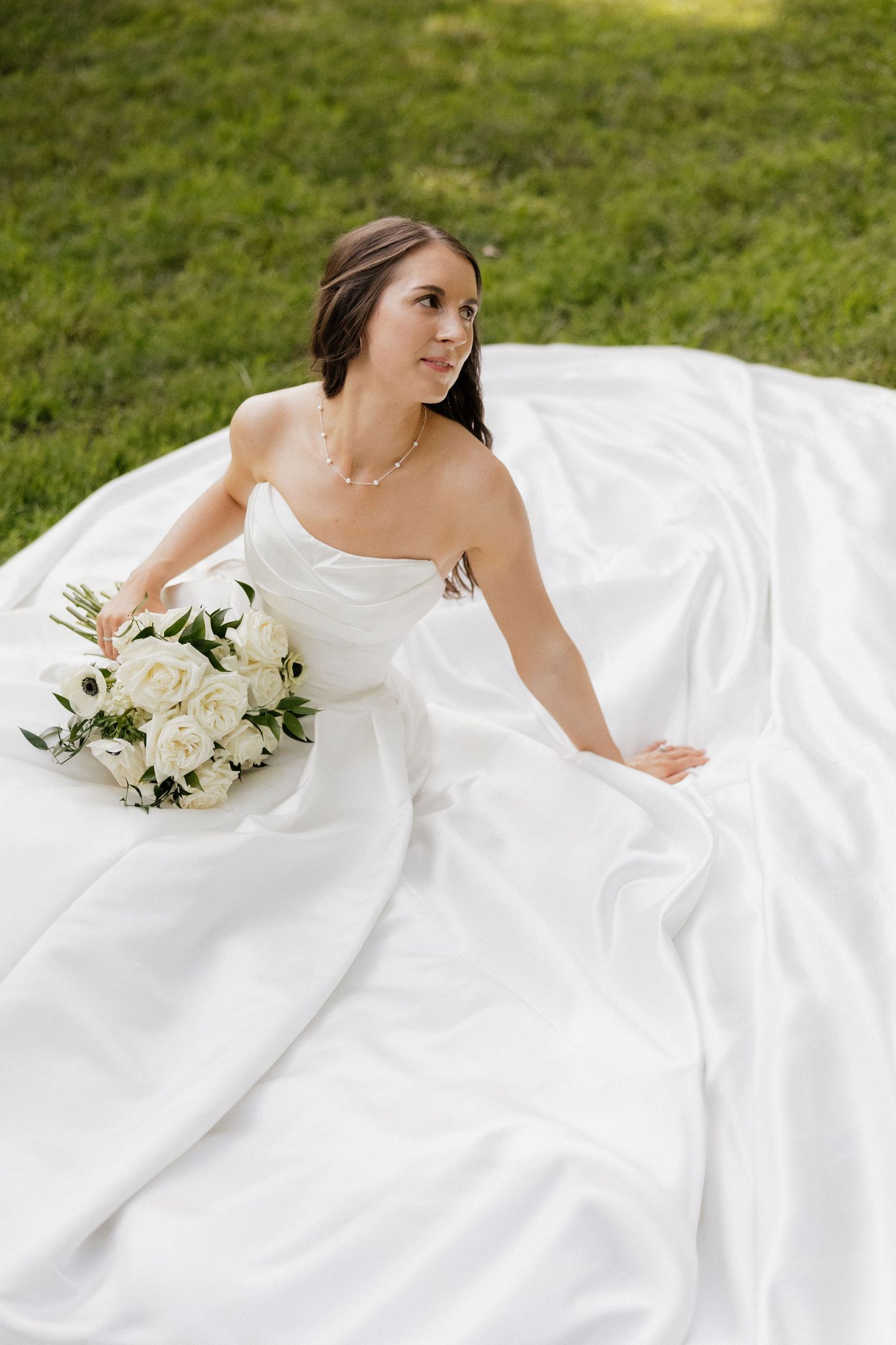 Bride in white strapless gown sitting on grass holding white rose bouquet, looking away gracefully.