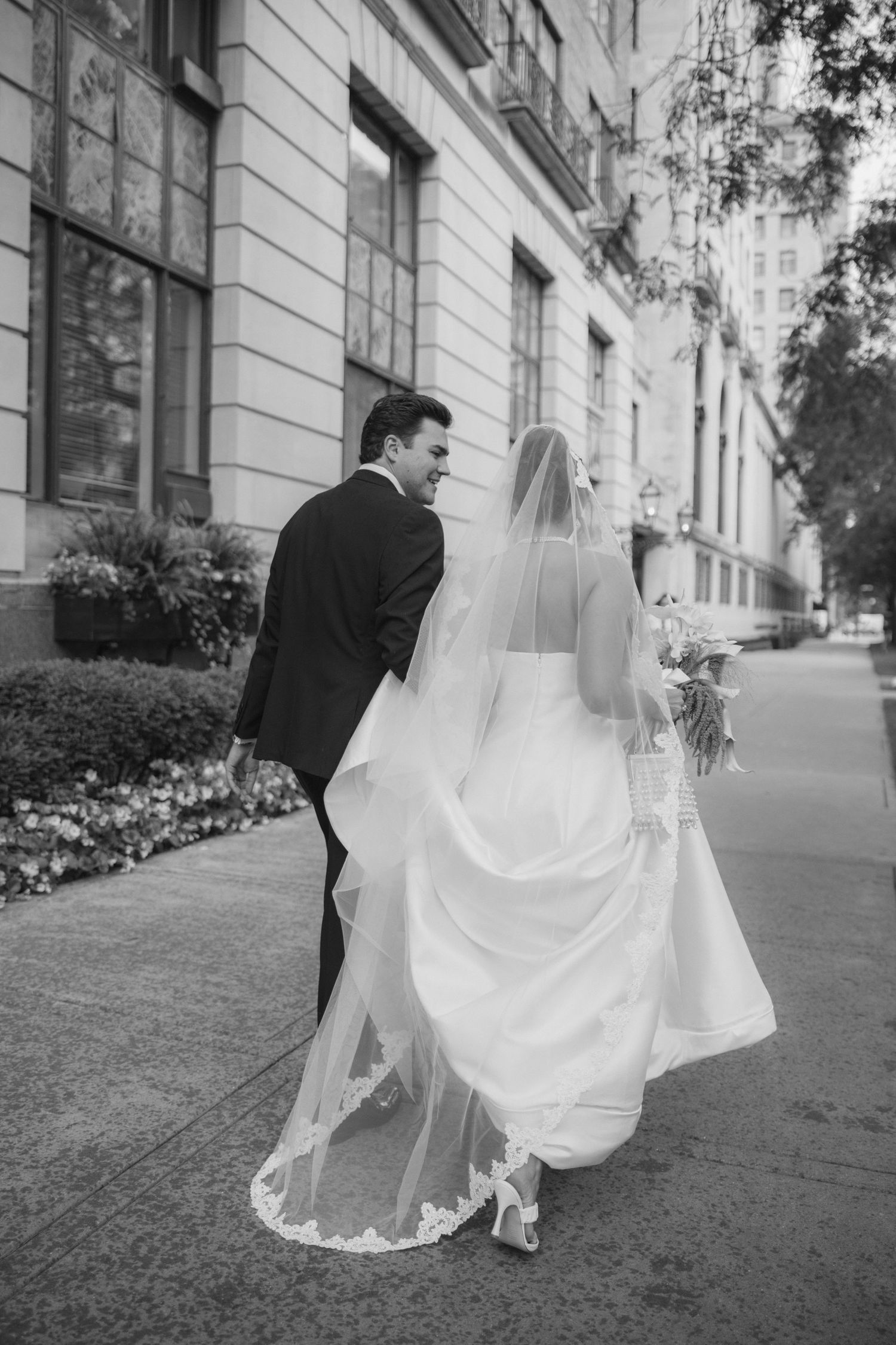 Black and white photo of bride and groom walking on city sidewalk, bride in flowing gown with lace veil.