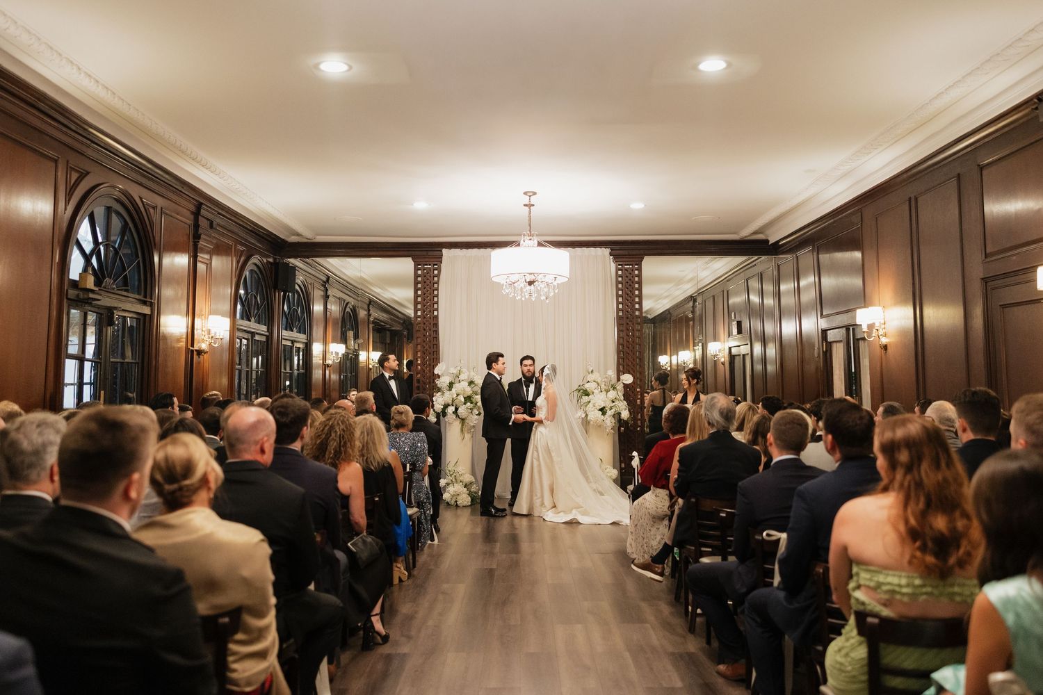 Elegant wedding ceremony in a wood-paneled ballroom with chandelier, guests seated as couple exchanges vows at floral altar.