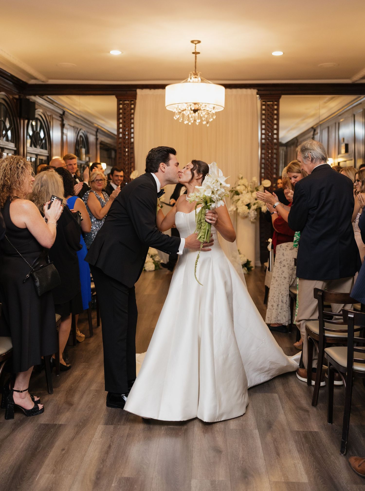 Bride and groom share first kiss as newlyweds while guests applaud in elegant wedding venue with chandelier.