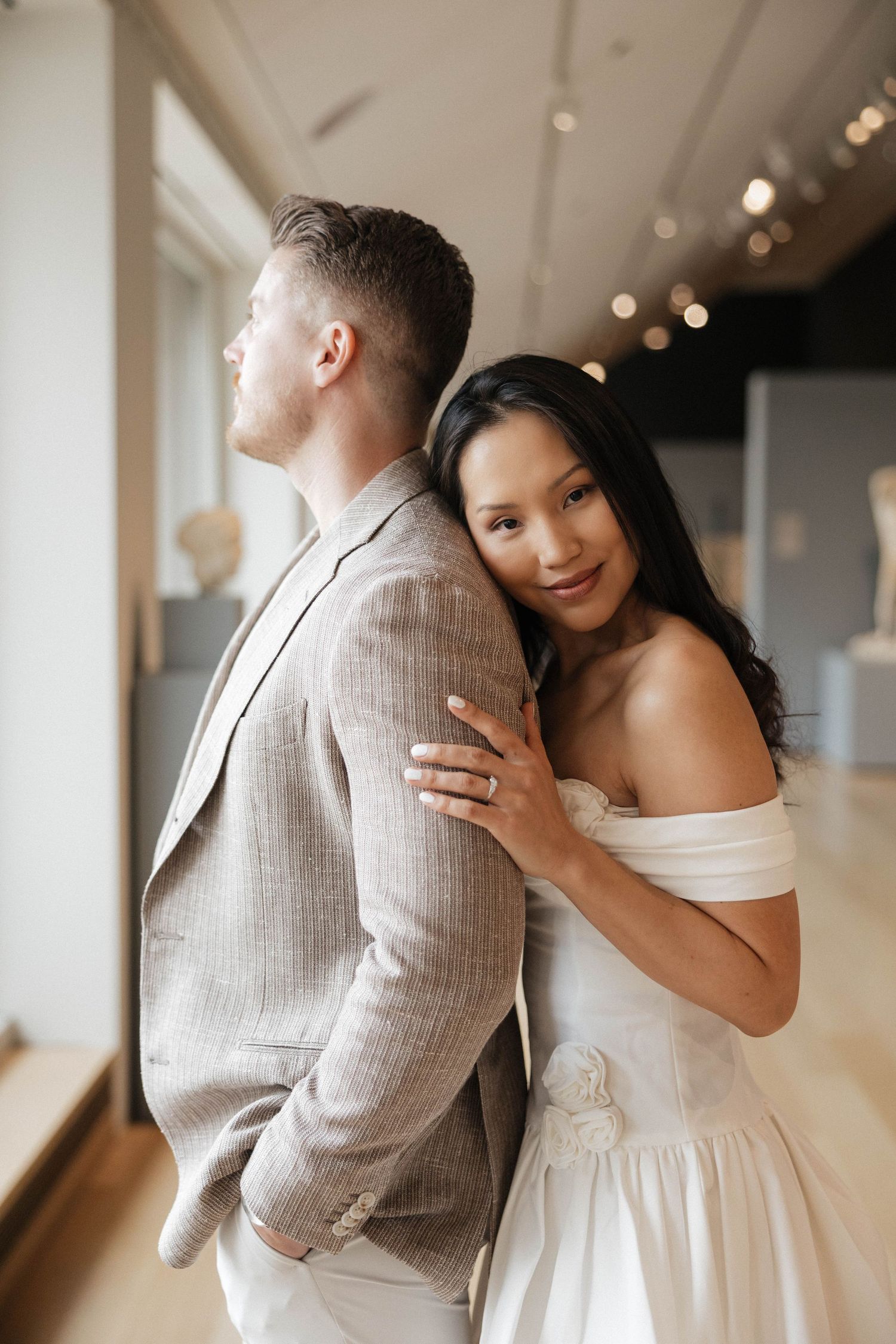 A bride in an off-shoulder white dress embraces a groom in a linen suit inside an elegant museum gallery.