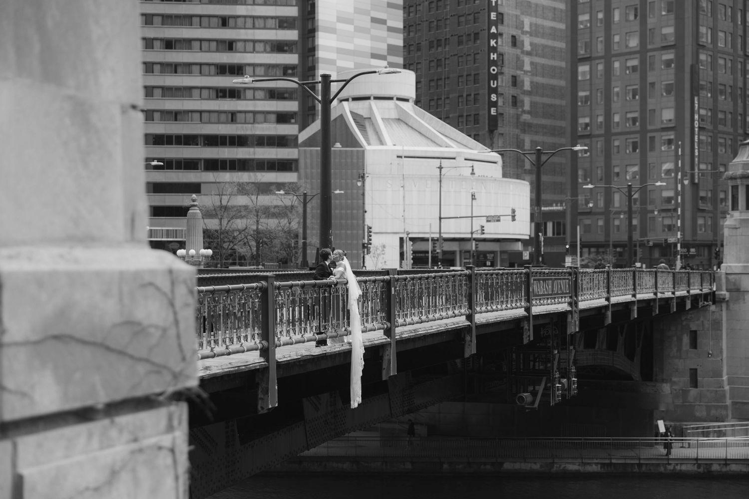 Black and white photo of a bridge with ornate railings spanning a river in a urban cityscape.