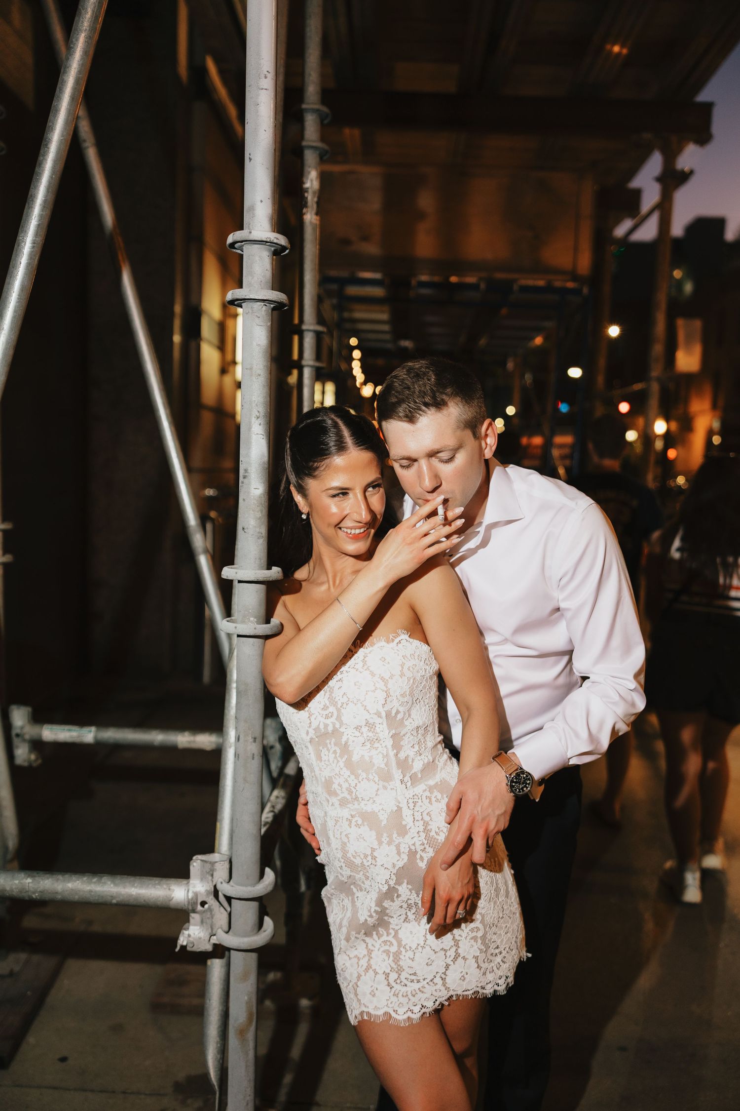 Bride in short lace dress and groom in white shirt share intimate moment on city street at night.