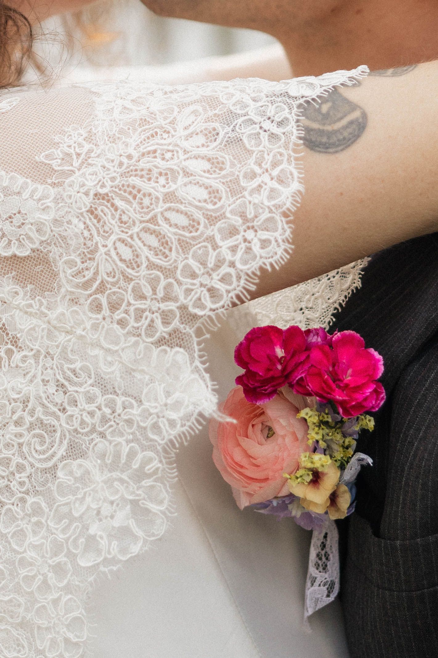 Close-up of bride in lace wedding dress embracing groom with pink floral boutonniere, tattoo visible on arm.