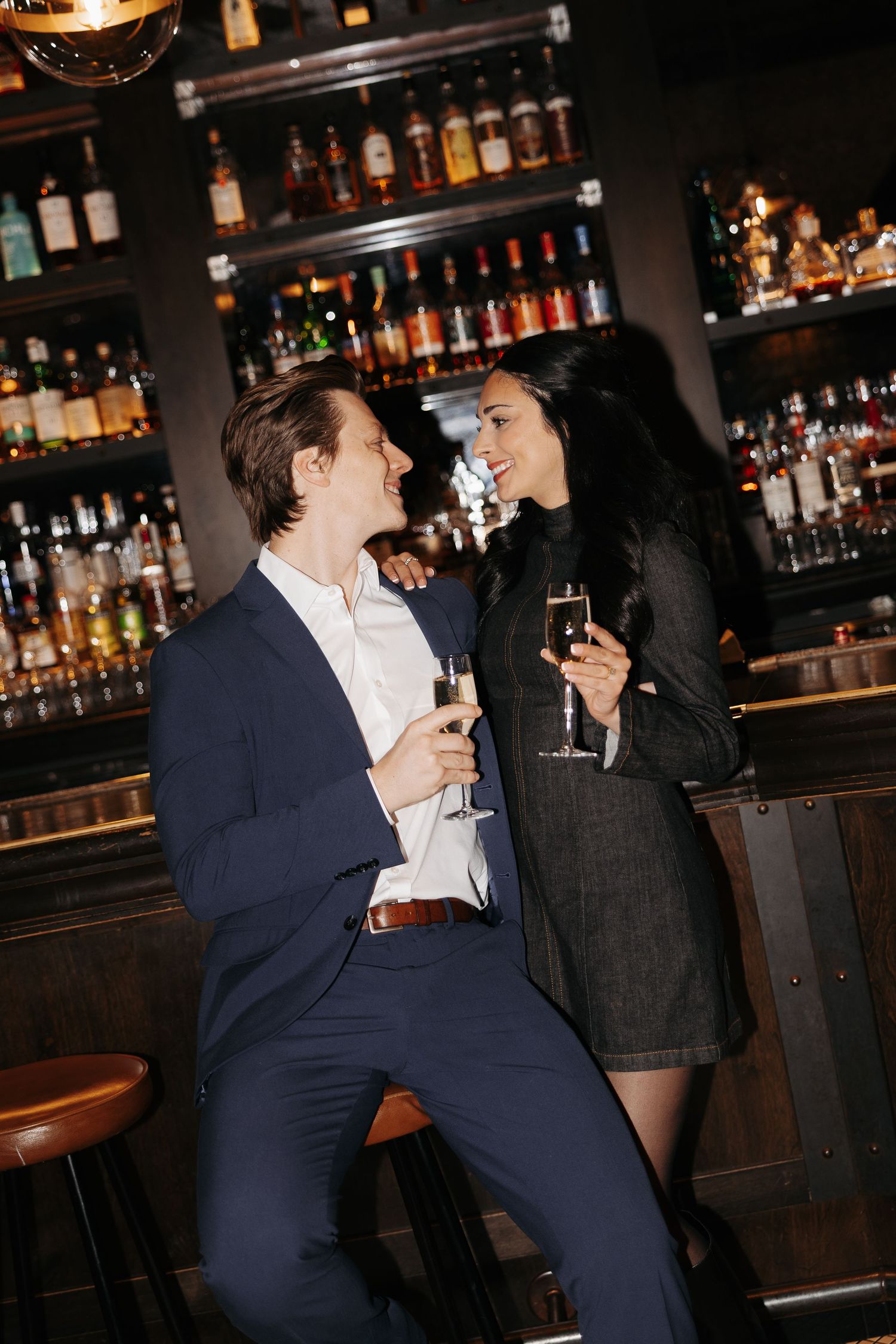 Two people share an intimate moment at a dimly lit bar with bottles displayed in the background.