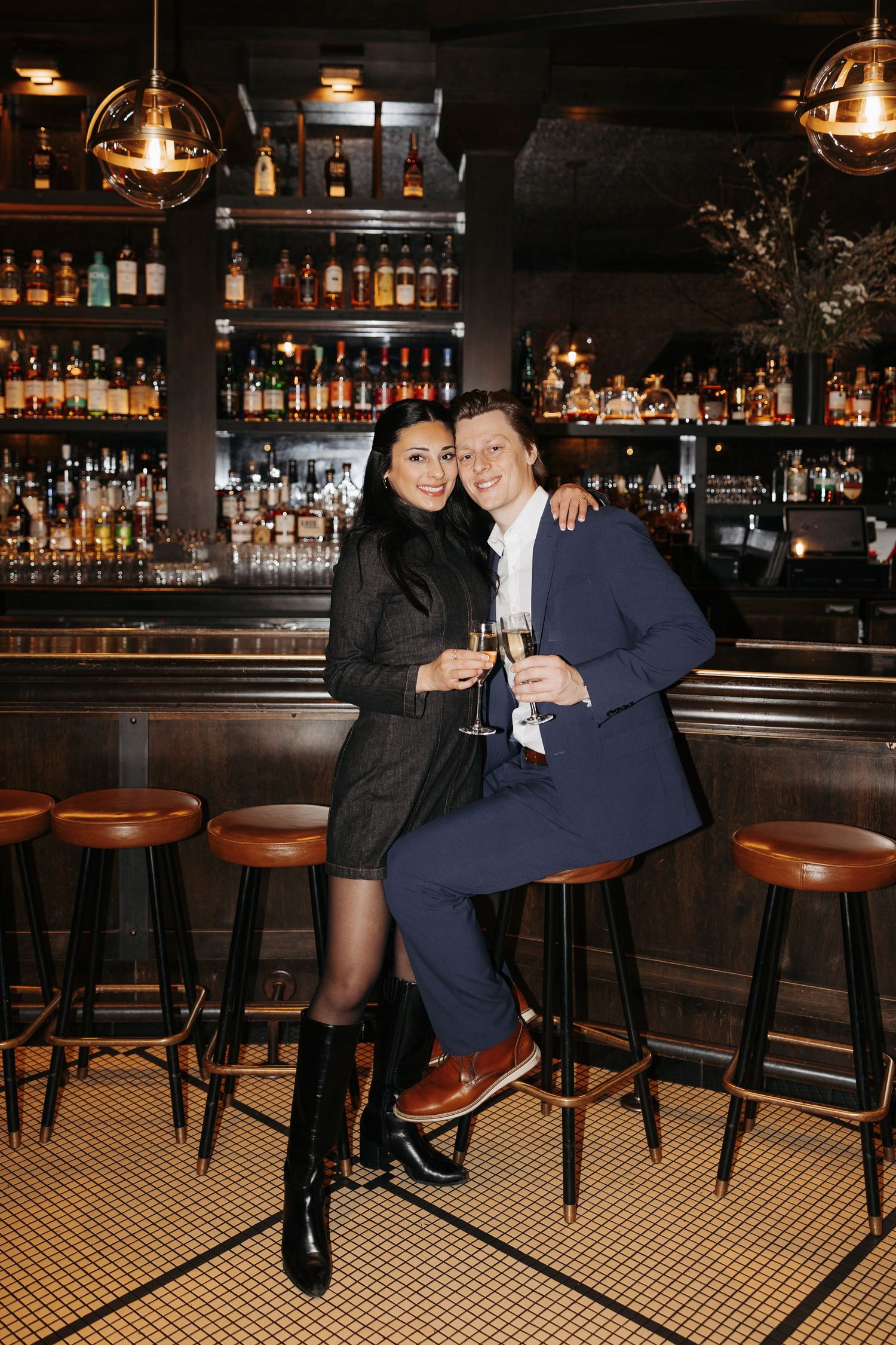 A couple enjoys drinks together at an elegant dark bar with wooden stools and illuminated bottle displays.