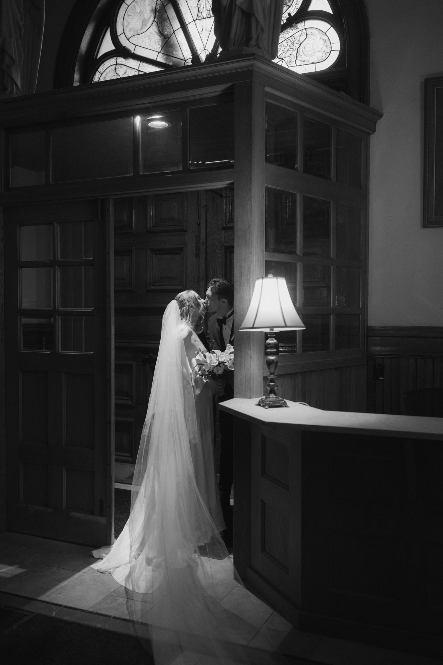 A bride in a flowing white gown and veil stands in a dimly lit hallway with vintage architectural details.