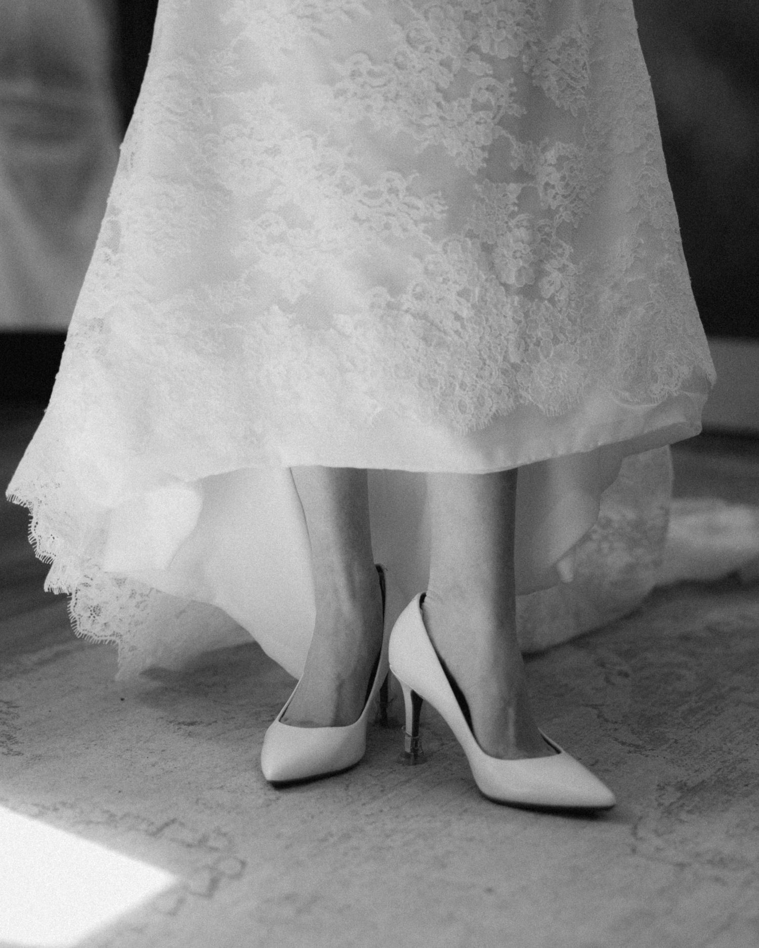 Close-up view of white wedding shoes and lace dress hem on a light colored floor.
