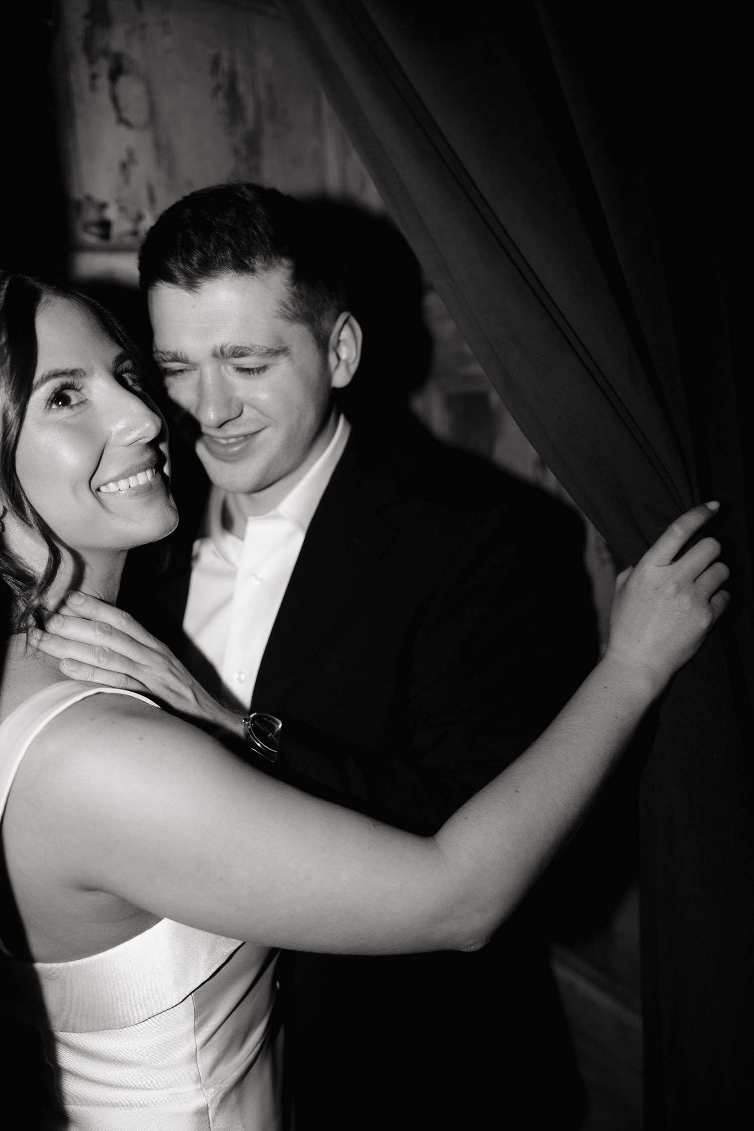 A black and white photo captures an intimate moment between a couple in formal attire near a curtain.