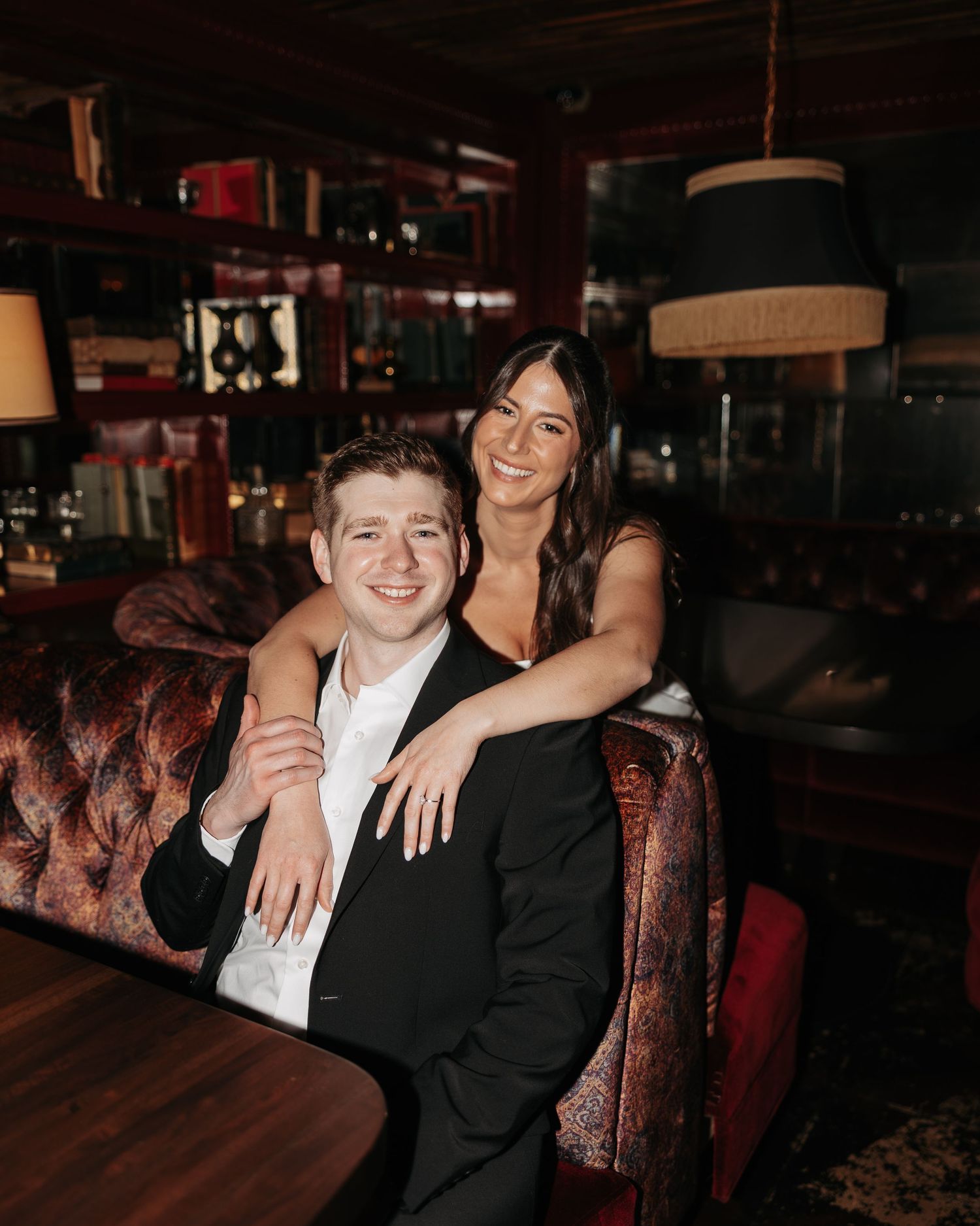 A couple in formal attire sits together on a vintage red velvet couch in a dimly lit venue.