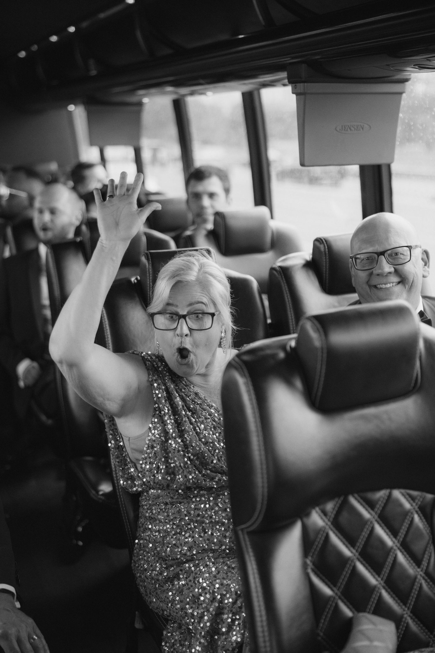 Excited woman in sequin dress raises hand on party bus, smiling bald man with glasses visible behind her.