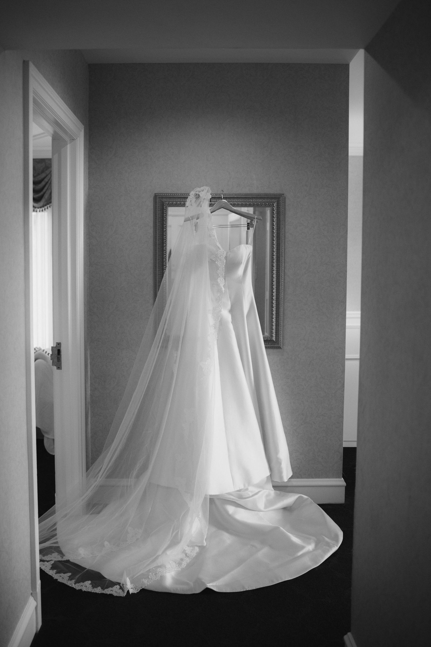 Black and white photo of an elegant wedding dress with veil hanging in a bridal suite before the ceremony.