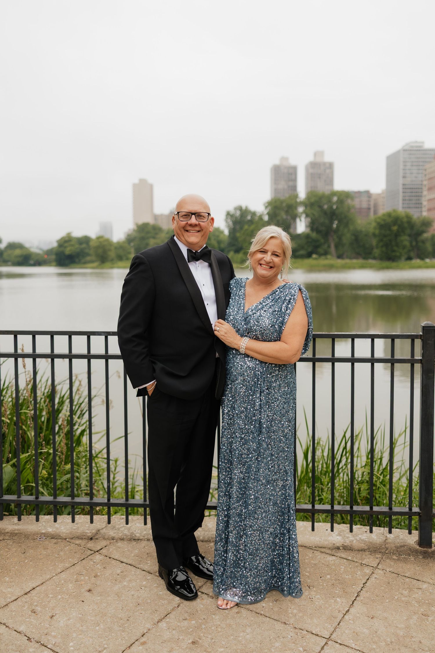 Elegant couple in formal attire posing by a lakeside railing with city skyline in background.