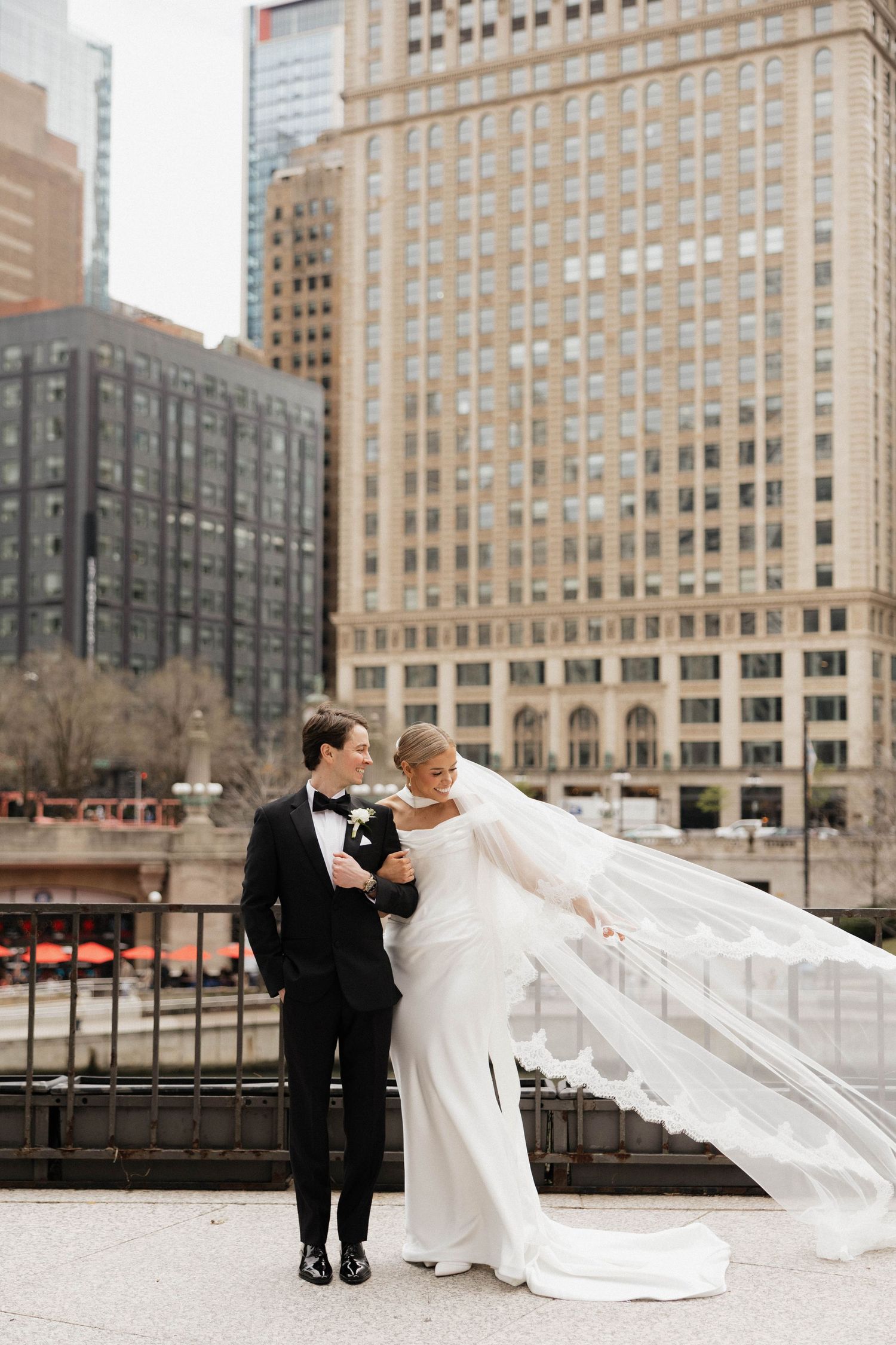 Bride in flowing white gown with veil and groom in black tuxedo pose on urban terrace with city buildings backdrop.