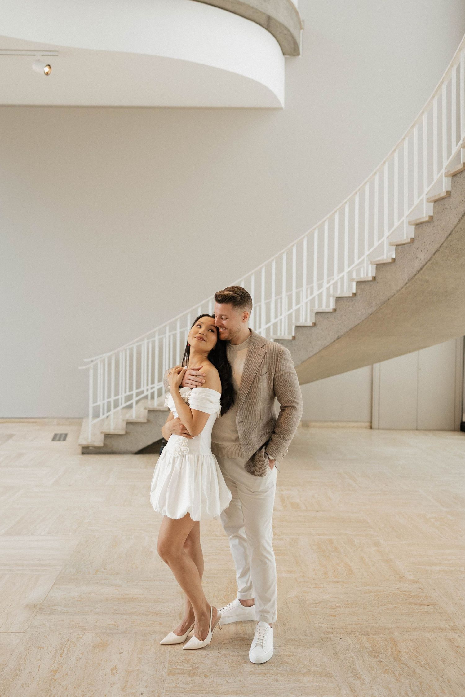 Elegant couple embracing in a modern white interior with a curved staircase, woman in white mini dress, man in beige suit.