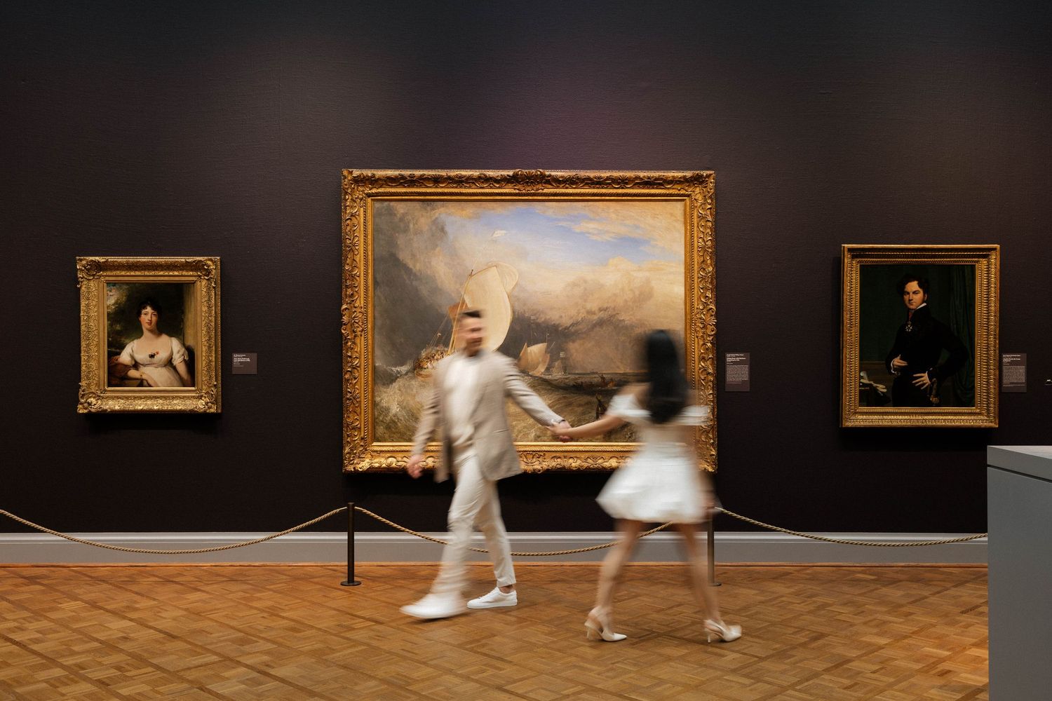 Two blurred figures dance through a museum gallery past ornate gold-framed paintings on dark walls.