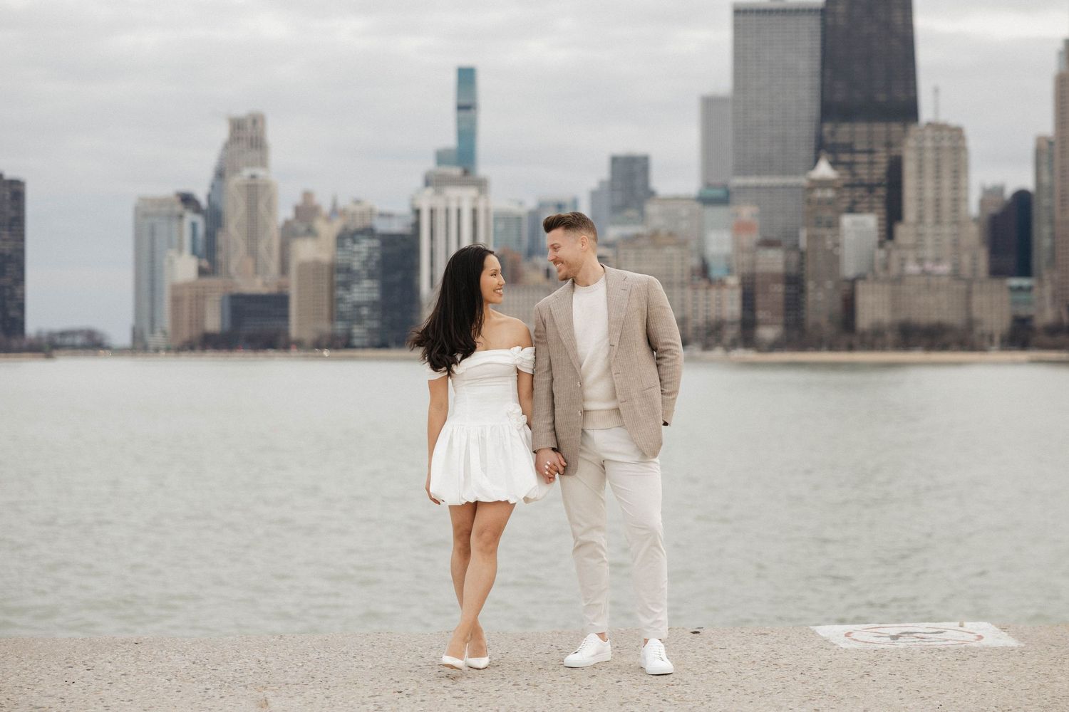 Couple holding hands and gazing at each other by Chicago skyline waterfront, dressed in white and tan outfits.