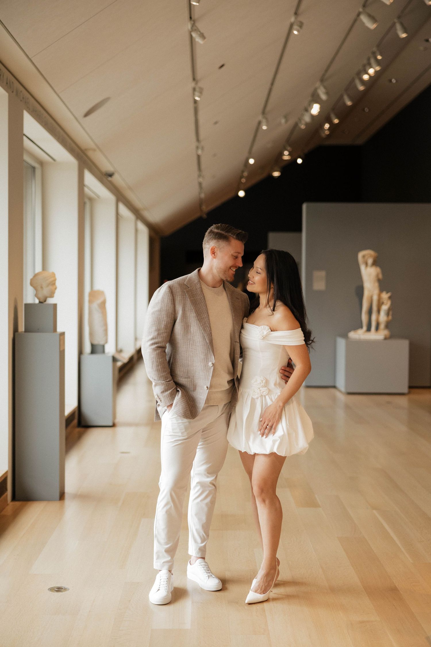 Couple in neutral tones sharing a romantic moment in a museum gallery with classical sculptures.