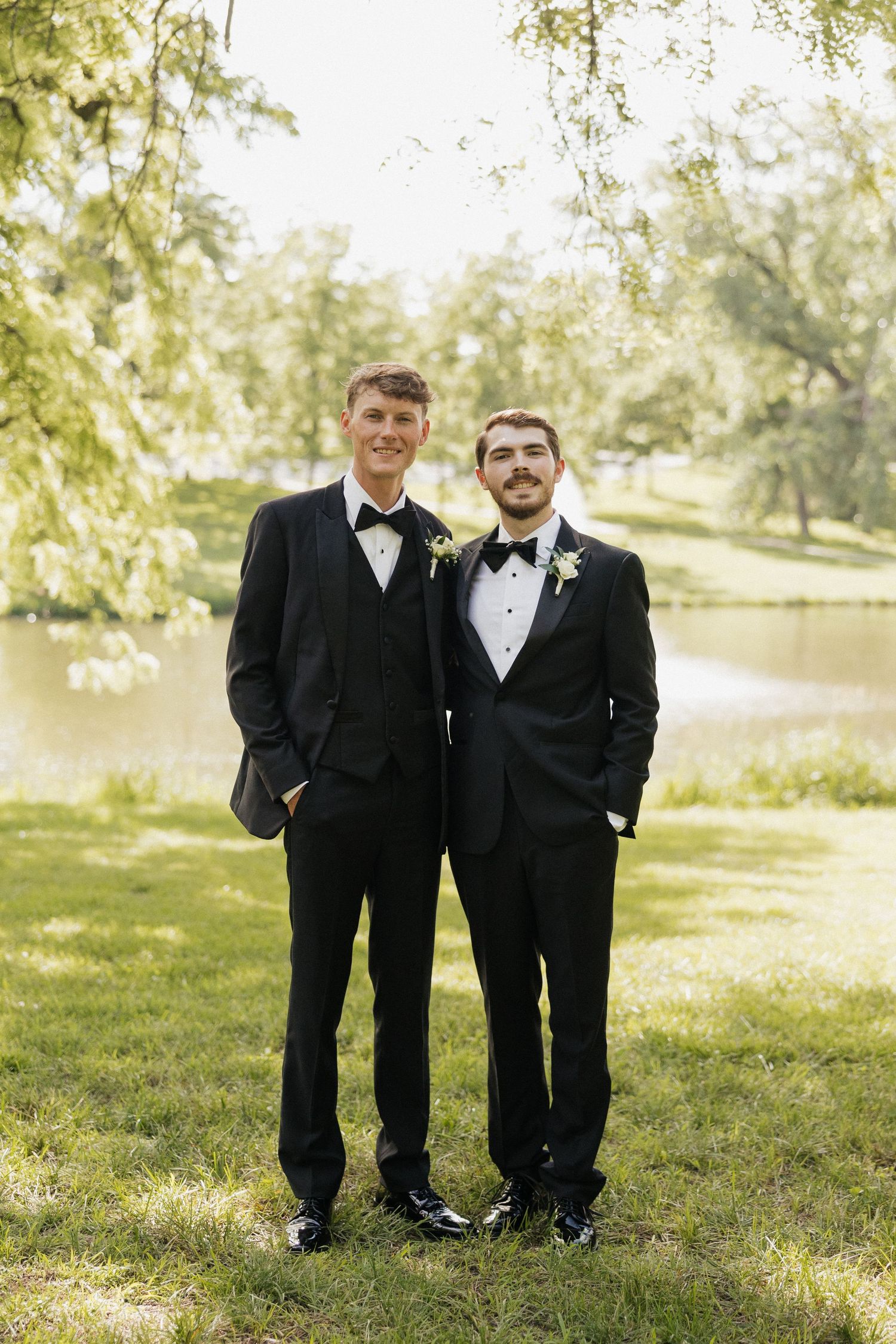 Two men in black tuxedos with bow ties and boutonnieres pose outdoors near a pond surrounded by lush green trees.