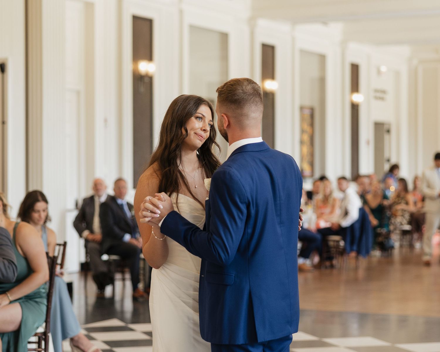 A couple shares their first dance at an elegant indoor wedding reception with guests watching from their seats.