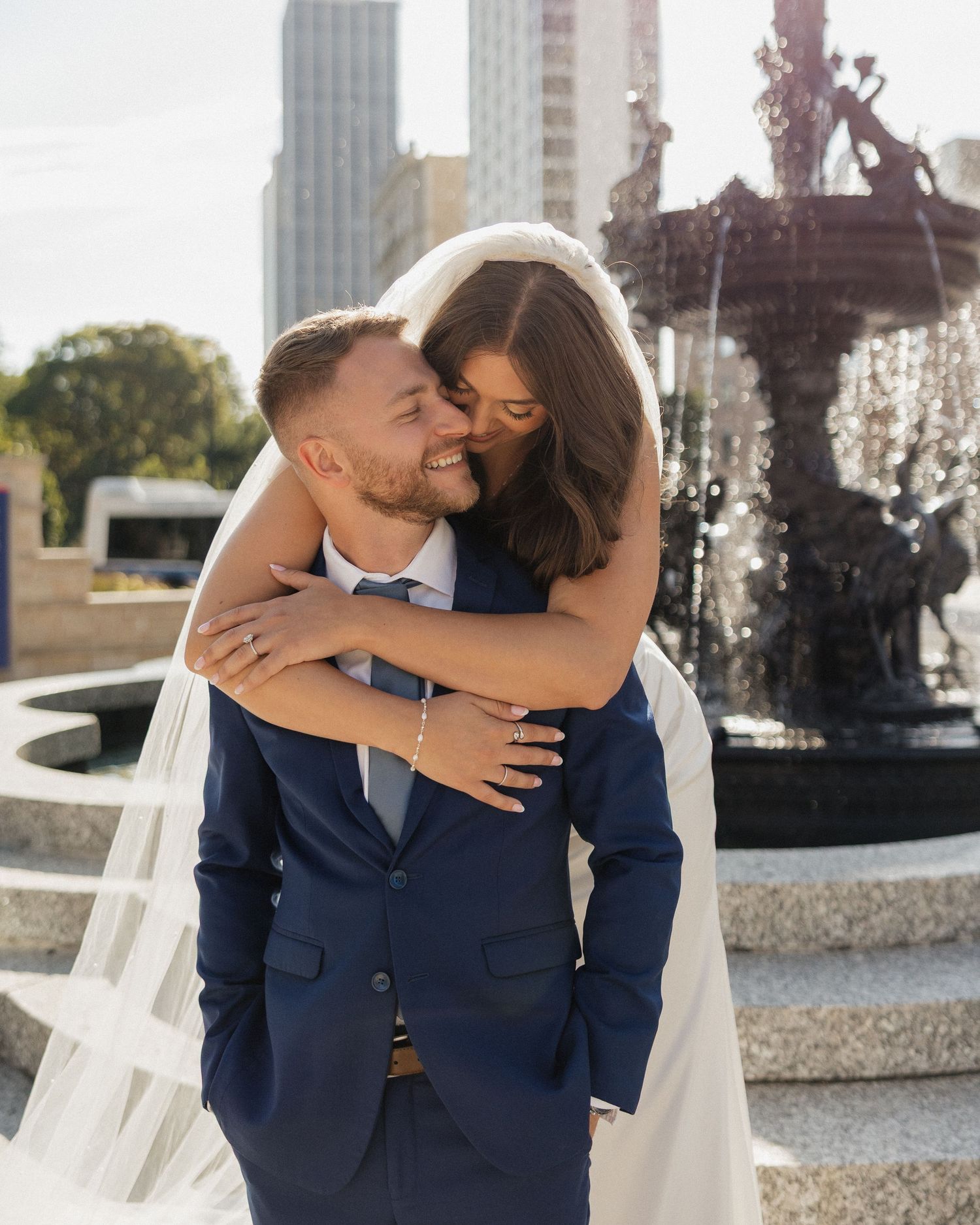 A couple in wedding attire embraces near an ornate fountain in an elegant outdoor setting.