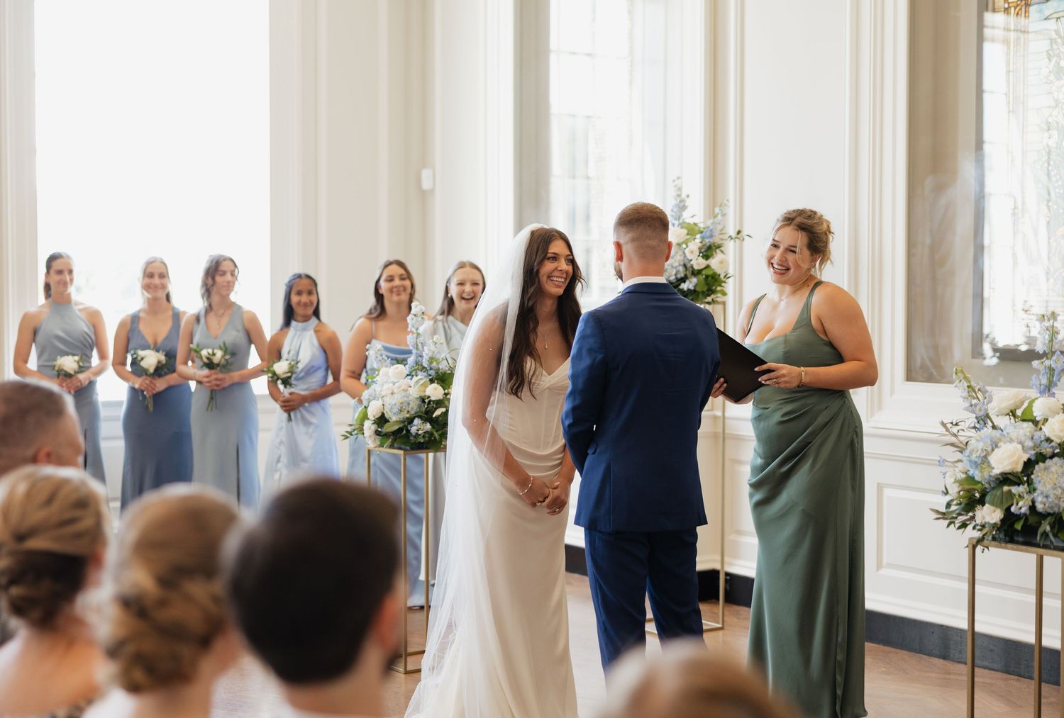 Wedding party members react with joy and excitement during an emotional moment in a bright indoor venue.