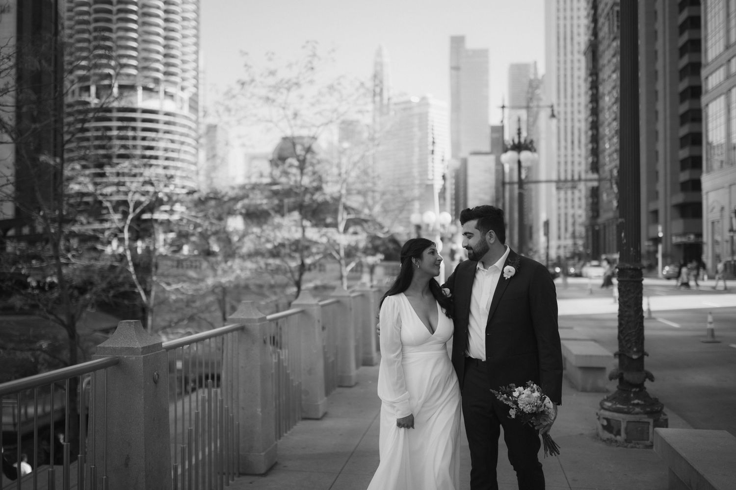 A couple in wedding attire poses together on an urban sidewalk with tall buildings in the background.